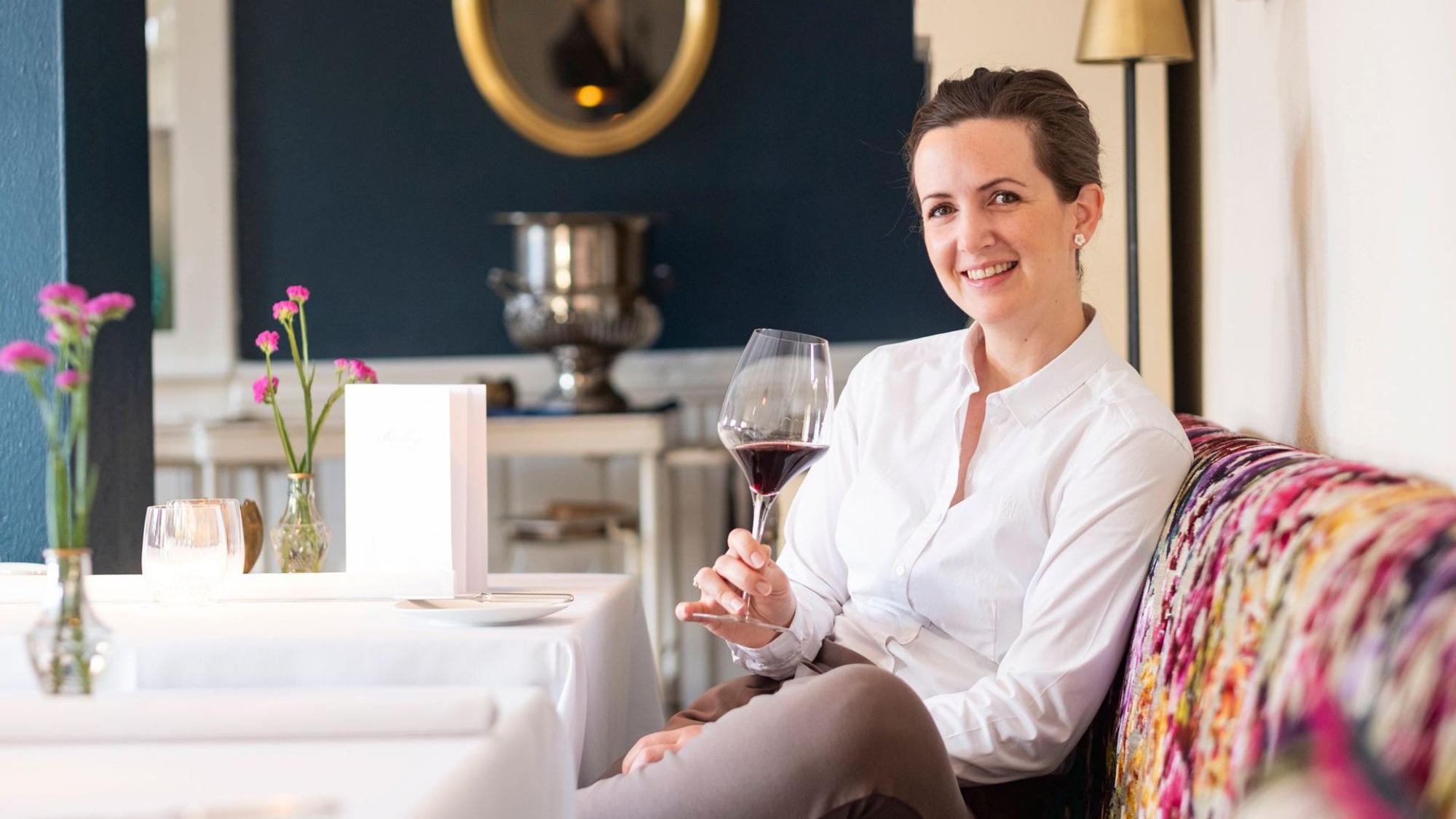 Katharina Röder sitzt in weißem Hemd mit einem Glas Wein in der Hand auf einer bunt gepolsterten Sitzbank an einem weiß gedeckten Tisch.