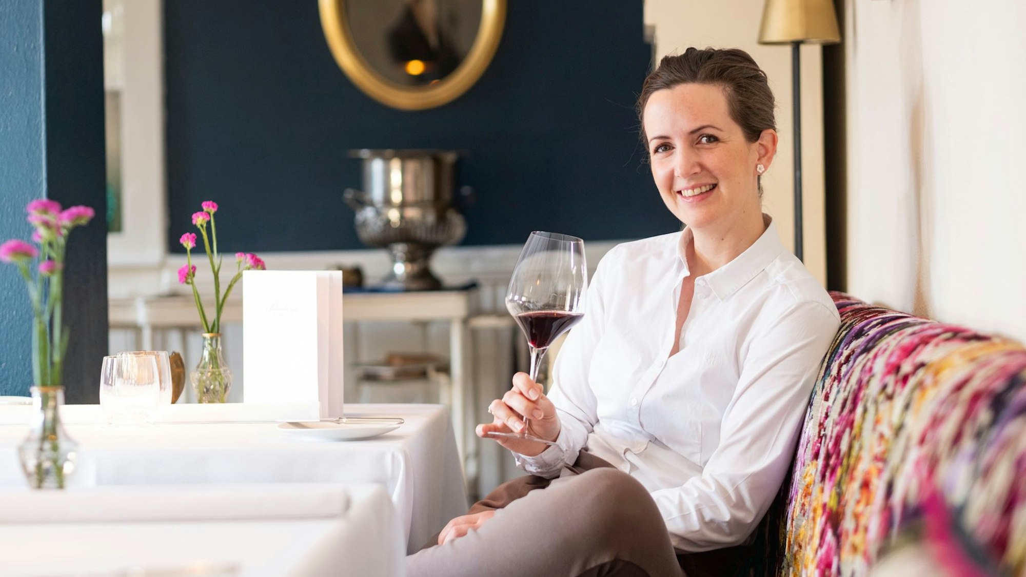 Katharina Röder sitzt in weißem Hemd mit einem Glas Wein in der Hand auf einer bunt gepolsterten Sitzbank an einem weiß gedeckten Tisch.