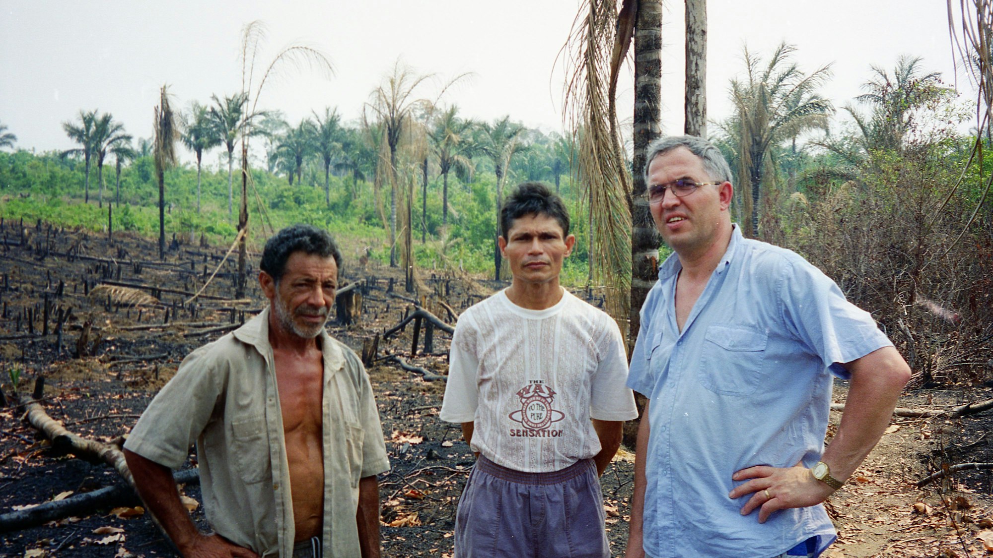 Walter Feckinghaus steht mit zwei Brasilianern in einem abgebrannten Areal des Amazonas. Im Hintergrund ist ein unbeschädigter Teil des Waldes zu sehen.