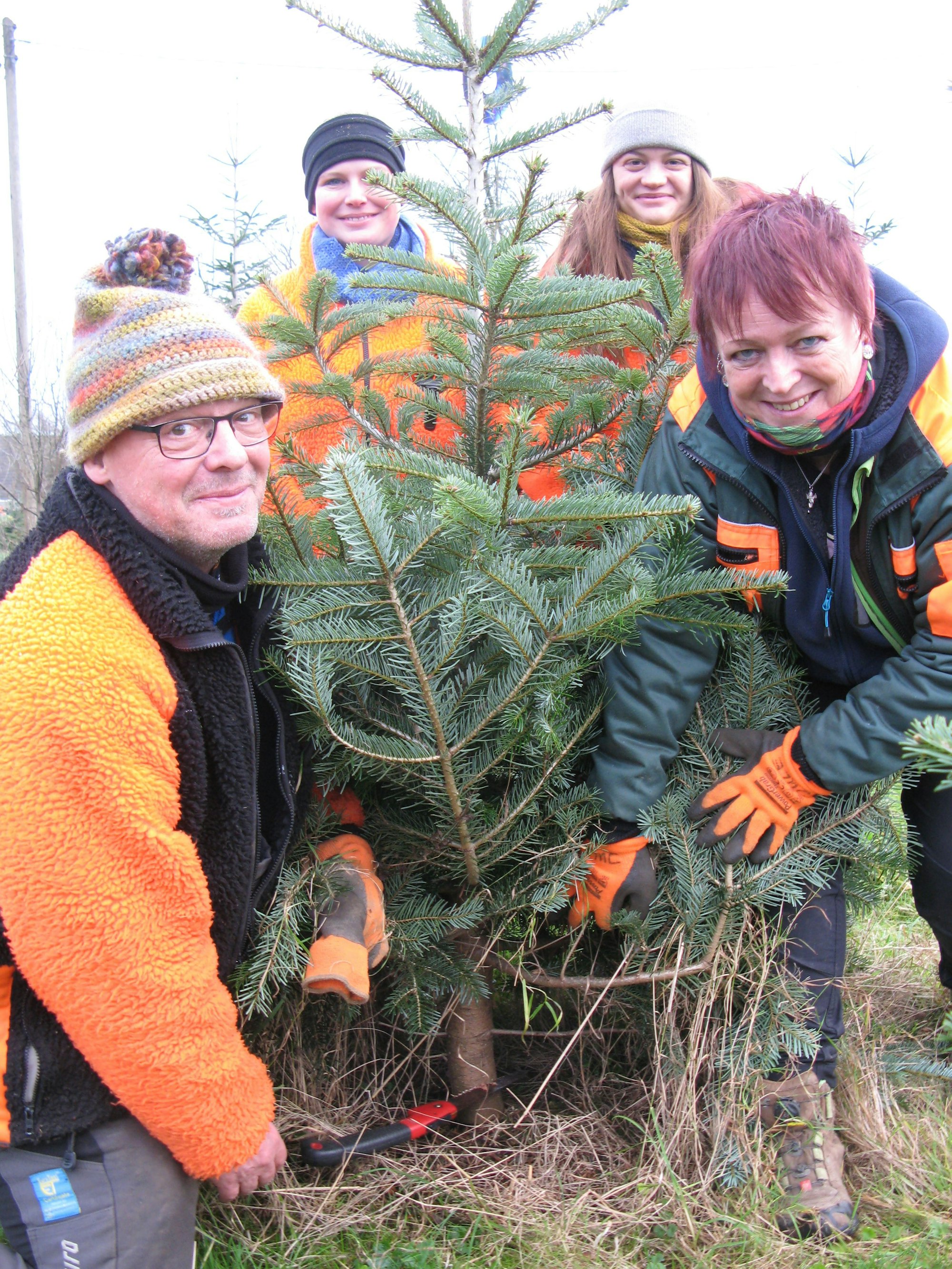 Ein Mann kniet neben dem Tannenbaum, den er absägen will. Seine Schwester und zwei Töchter stehen neben und hinter dem Baum, um zu helfen.