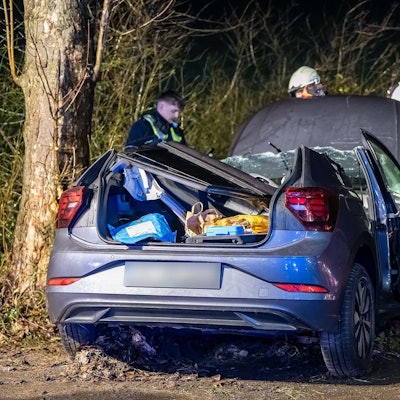 Ein Unfallwagen nach einem Aufprall gegen einen Baum. Mehrere Feuerwehrleute stehen um den Wagen herum.