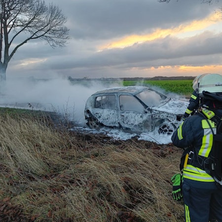 Ein Feuerwehrmann steht vor einem ausgebrannten, qualmenden Auto.