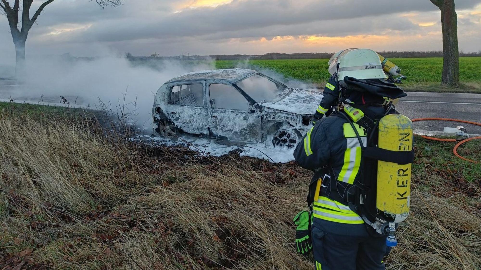 Ein Feuerwehrmann steht vor einem ausgebrannten, qualmenden Auto.