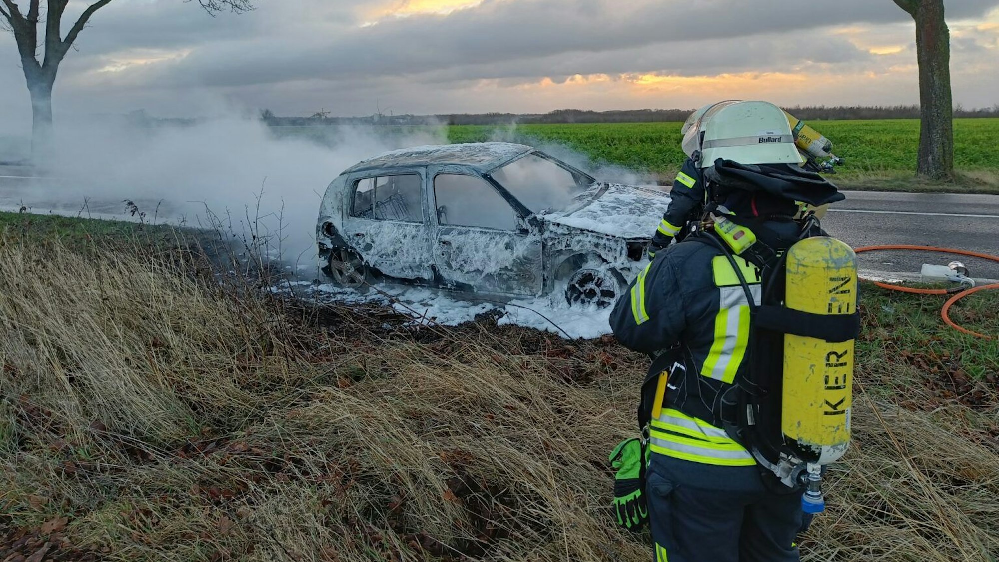 Ein Feuerwehrmann steht vor einem ausgebrannten, qualmenden Auto.