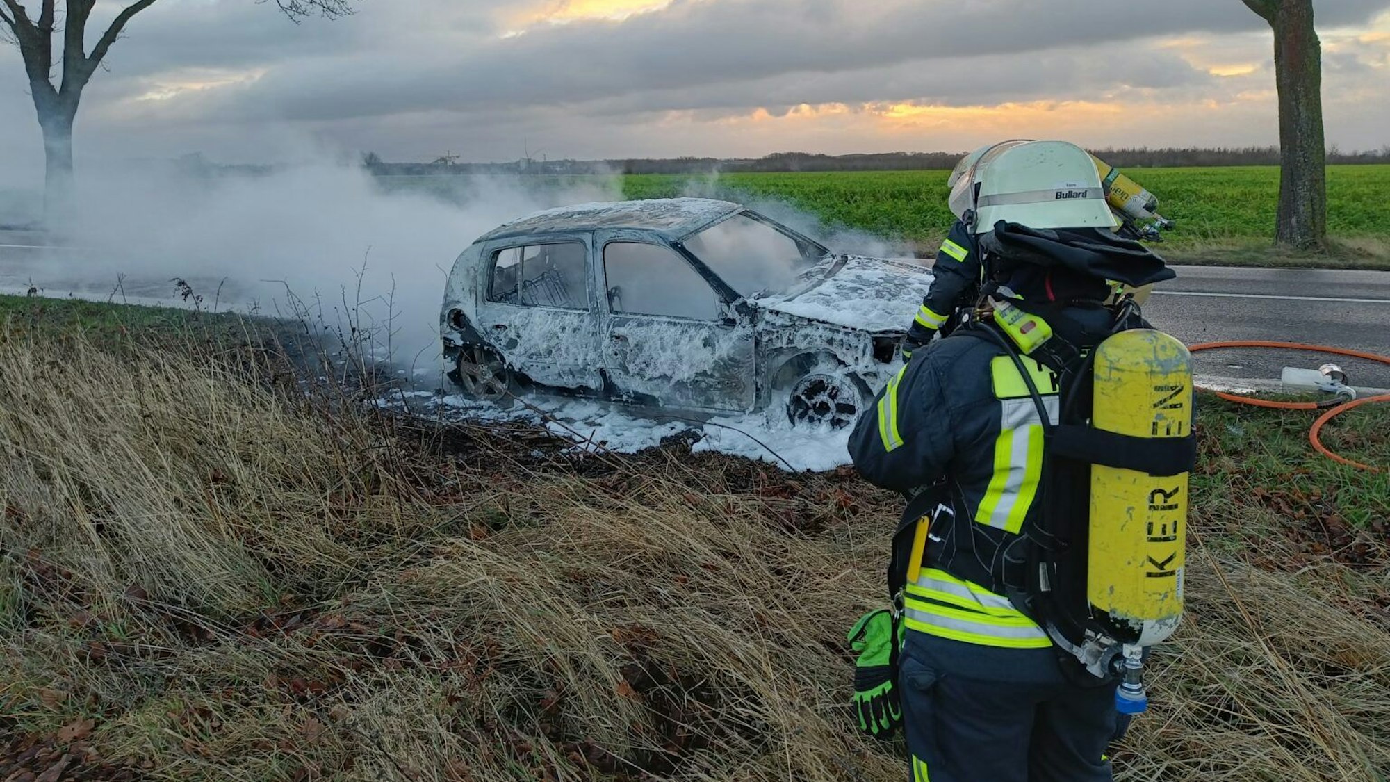 Ein Feuerwehrmann steht vor einem ausgebrannten, qualmenden Auto.