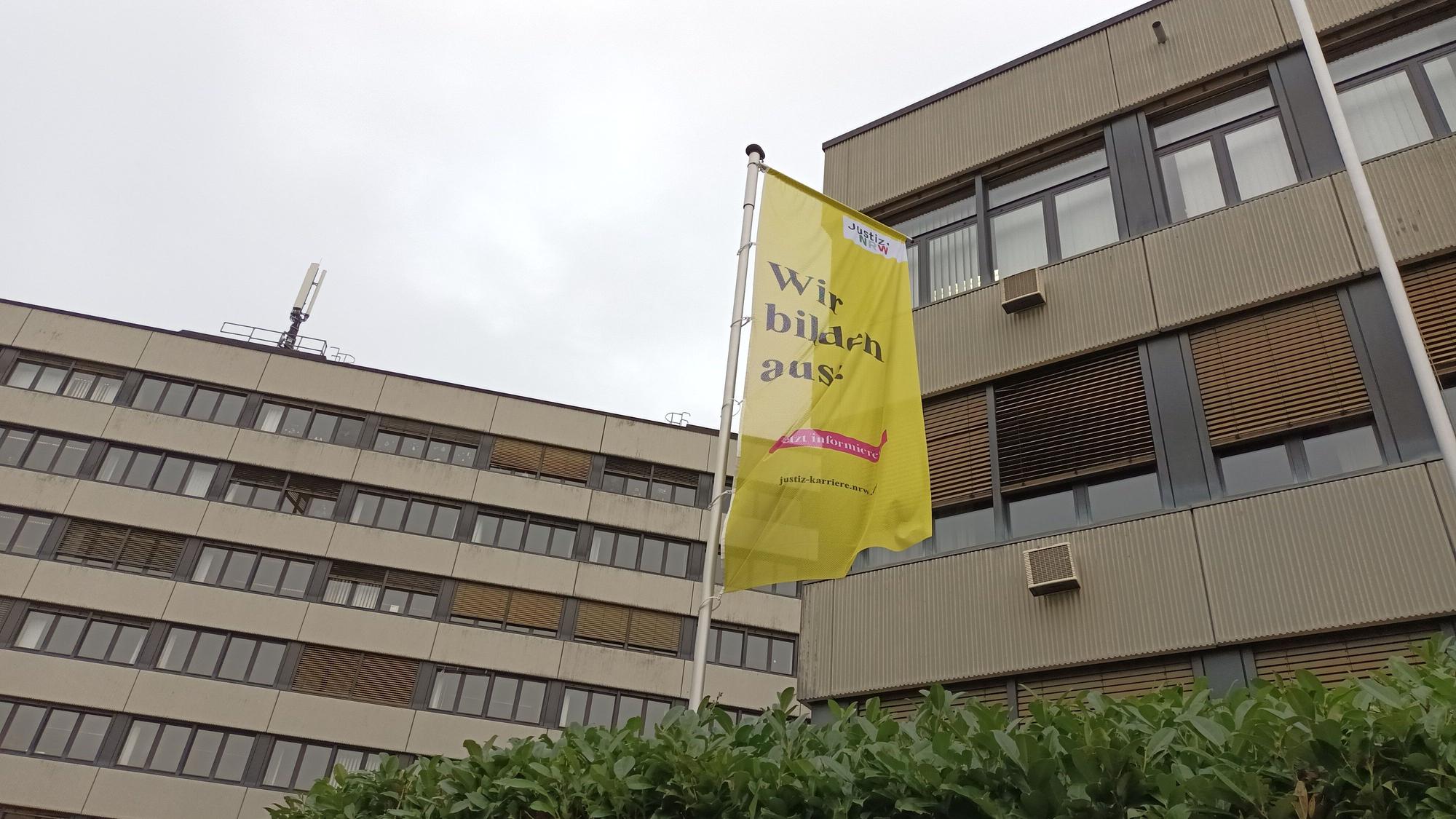 Das Bild zeigt die Fassade des Amtsgerichts in Siegburg mit einer Hecke und einer gelben Flagge.