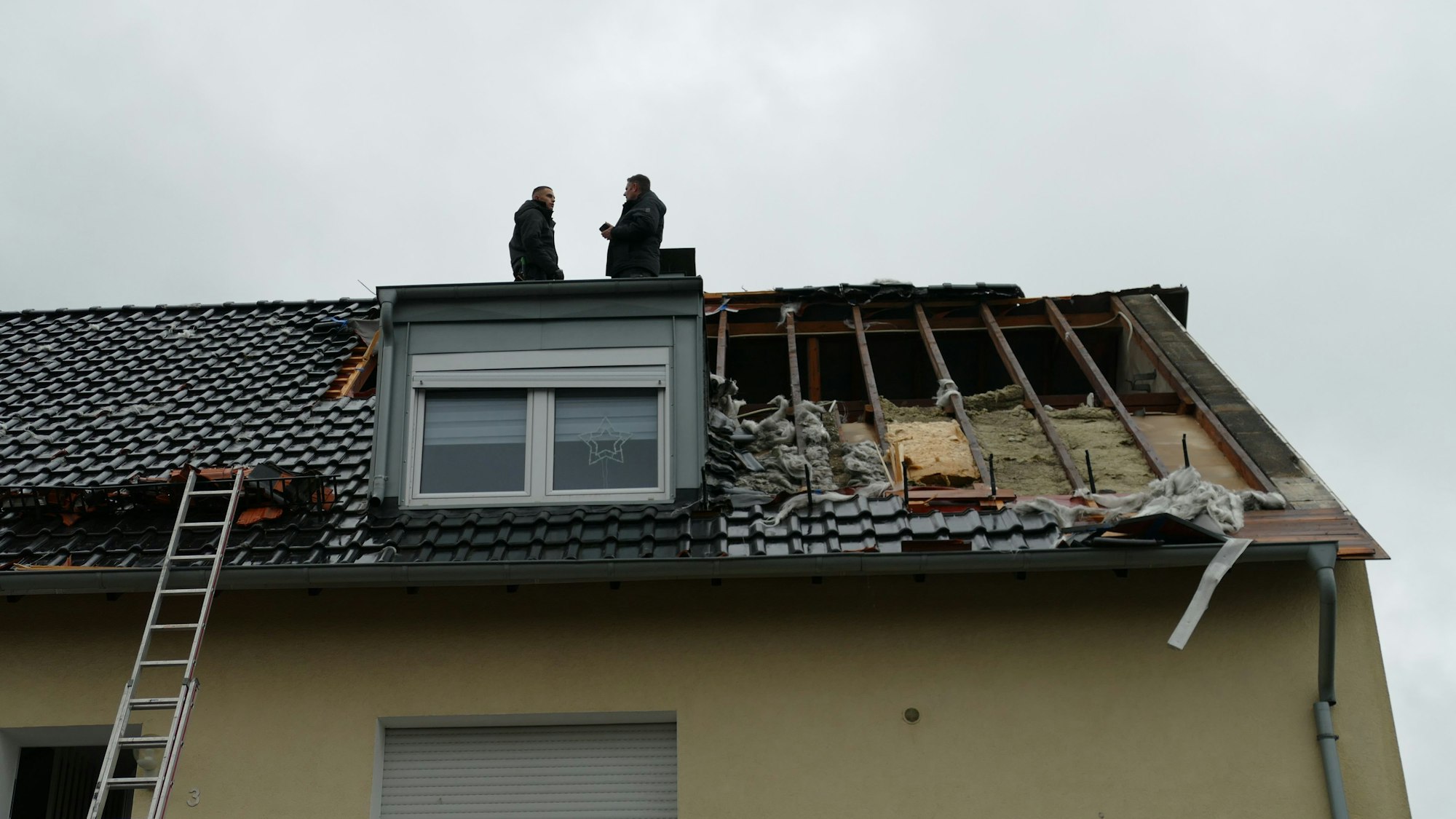 Zwei Männer stehen auf einem Dach, dessen Schindeln abgedeckt sind, große Löcher klaffen zwischen den Dachbalken.