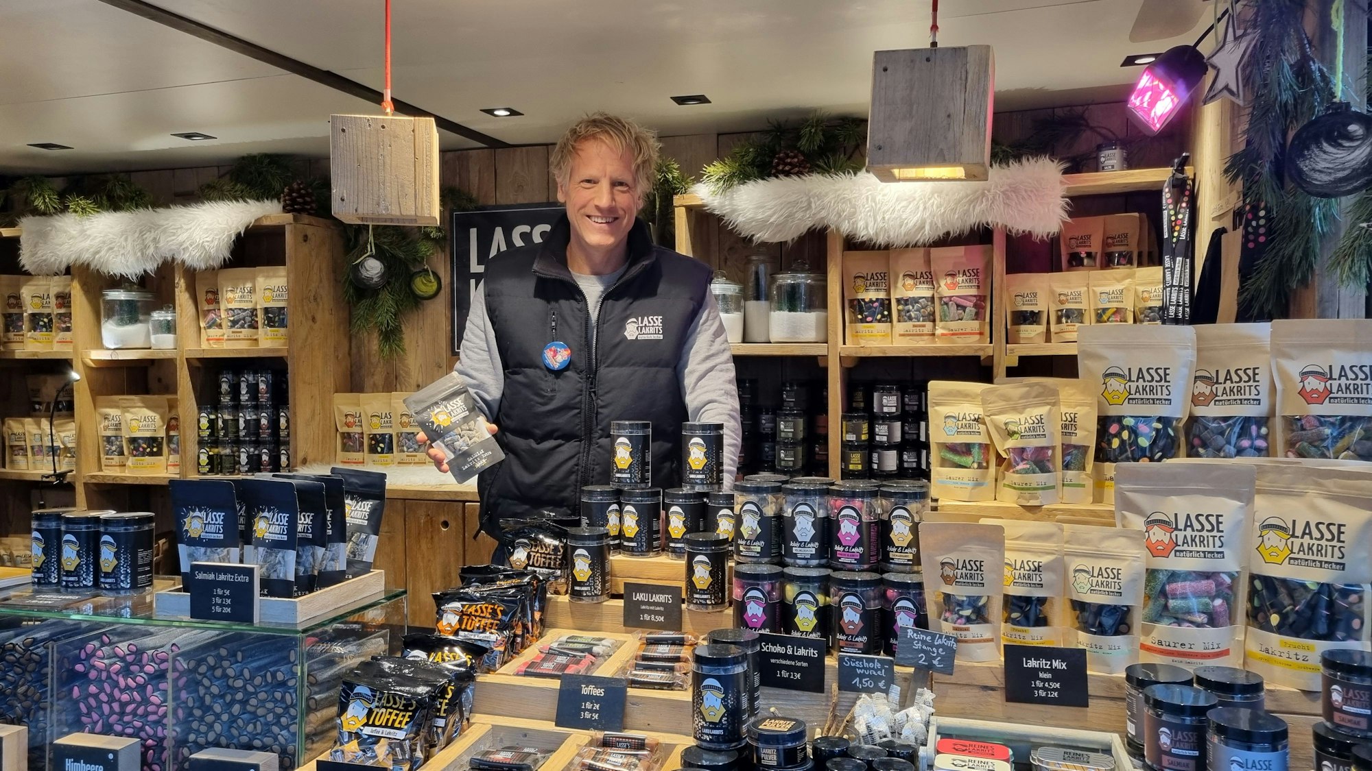 Zu sehen ist der hochgewachsene Firmengründer in einem Stand auf einem Weihnachtsmarkt. Um ihn herum Gläser, Tüten und Auslagen mit Lakritzspezialitäten in unterschiedlichsten Varianten.