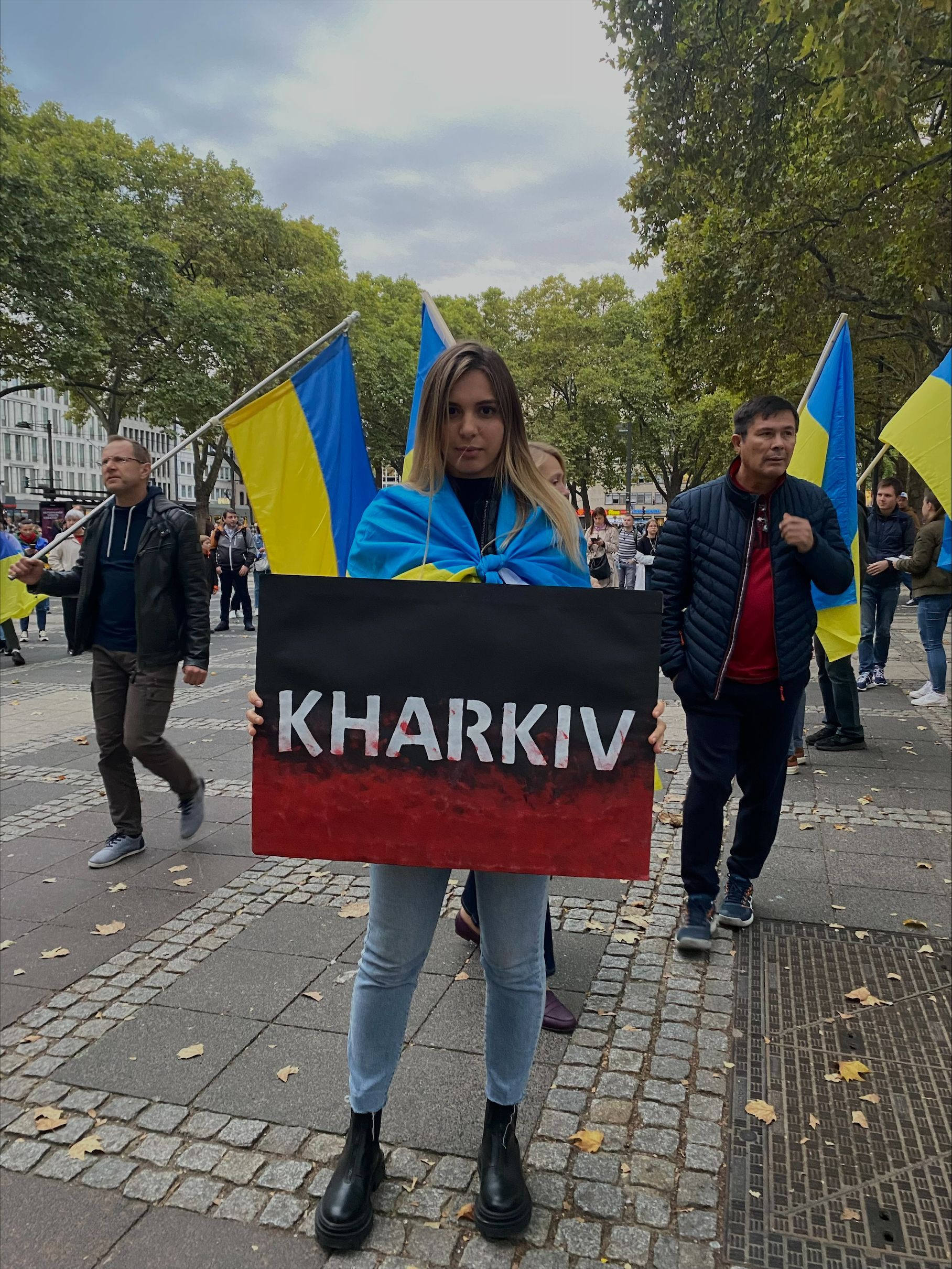 Lisa hält ein Schild, auf dem Charkiw steht und ist in eine Flagge der Ukraine gewickelt.