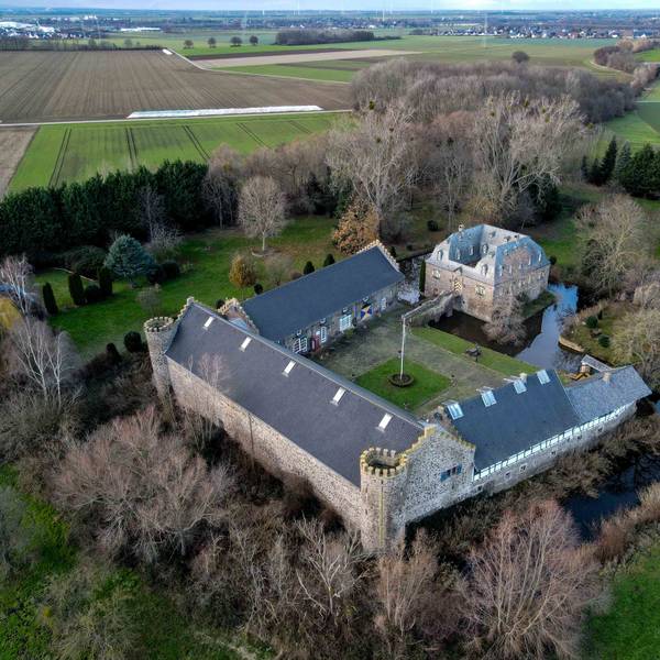 Das Foto zeigt das Burg-Ensemble aus der Luft. Im Vergleich zu den einen Hof umgebenden Wirtschaftsflügeln mit den beiden runden Türmen der Vorburg ist das über eine Zugbrücke zu erreichende Herrenhaus relativ klein. Umgeben ist die Anlage von Wassergräben und einem Park.