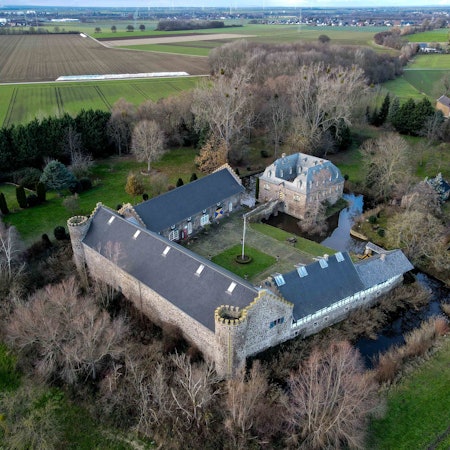 Das Foto zeigt das Burg-Ensemble aus der Luft. Im Vergleich zu den einen Hof umgebenden Wirtschaftsflügeln mit den beiden runden Türmen der Vorburg ist das über eine Zugbrücke zu erreichende Herrenhaus relativ klein. Umgeben ist die Anlage von Wassergräben und einem Park.