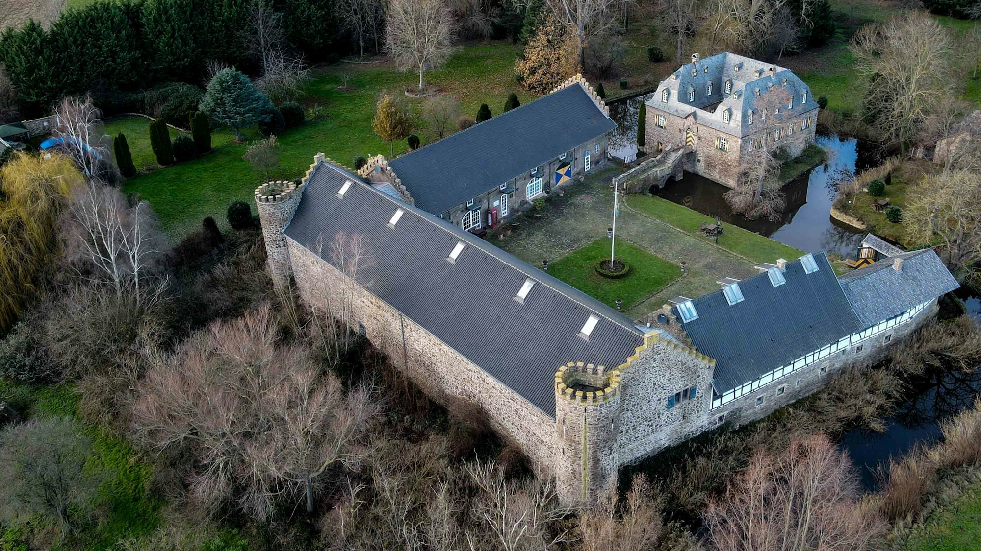 Das Foto zeigt das Burg-Ensemble aus der Luft. Im Vergleich zu den einen Hof umgebenden Wirtschaftsflügeln mit den beiden runden Türmen der Vorburg ist das über eine Zugbrücke zu erreichende Herrenhaus relativ klein. Umgeben ist die Anlage von Wassergräben und einem Park.