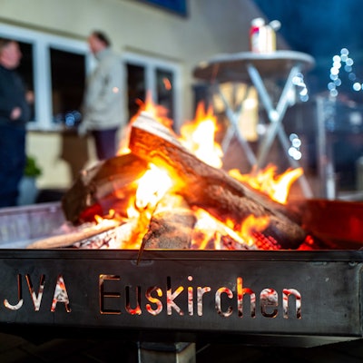 Das Bild zeigt eine Feuerschale in der JVA. Im Hintergrund stehen zwei Männer und unterhalten sich.