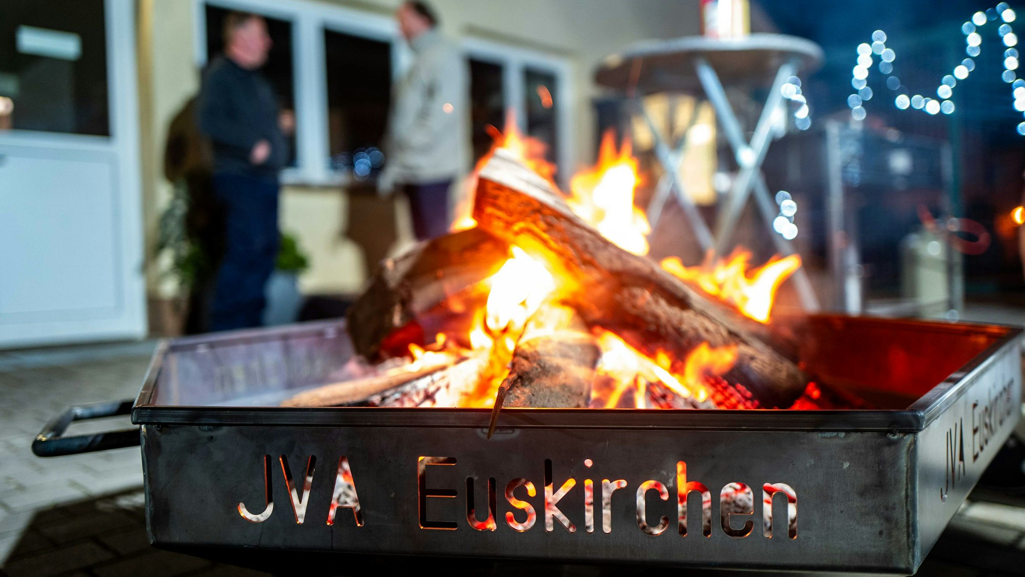Das Bild zeigt eine Feuerschale in der JVA. Im Hintergrund stehen zwei Männer und unterhalten sich.