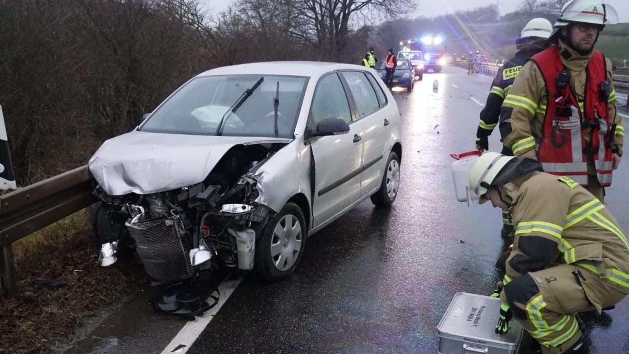 Ein nach einem Unfall beschädigtes Auto steht an einer Leitplanke, rechts sind drei Feuerwehrleute zu sehen.