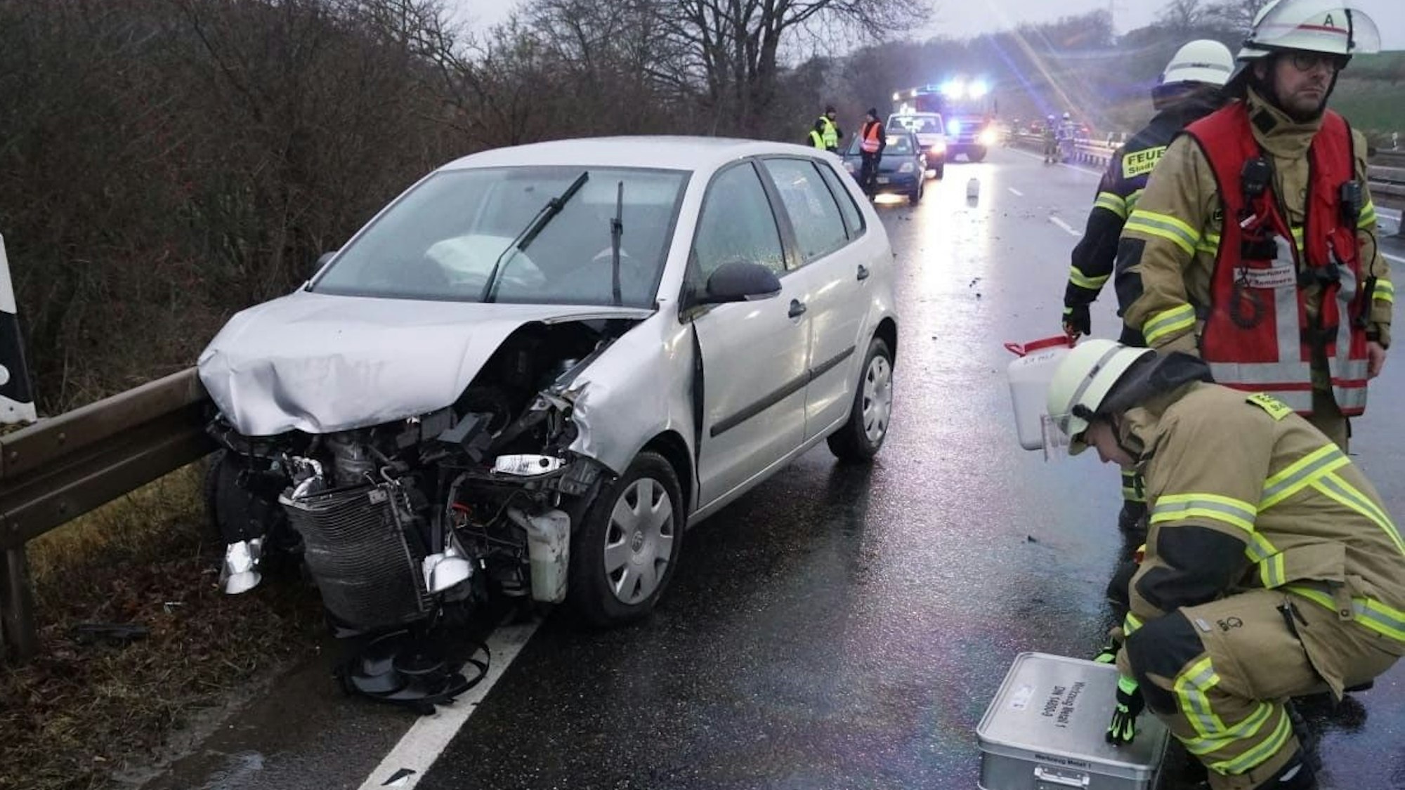 Ein nach einem Unfall beschädigtes Auto steht an einer Leitplanke, rechts sind drei Feuerwehrleute zu sehen.