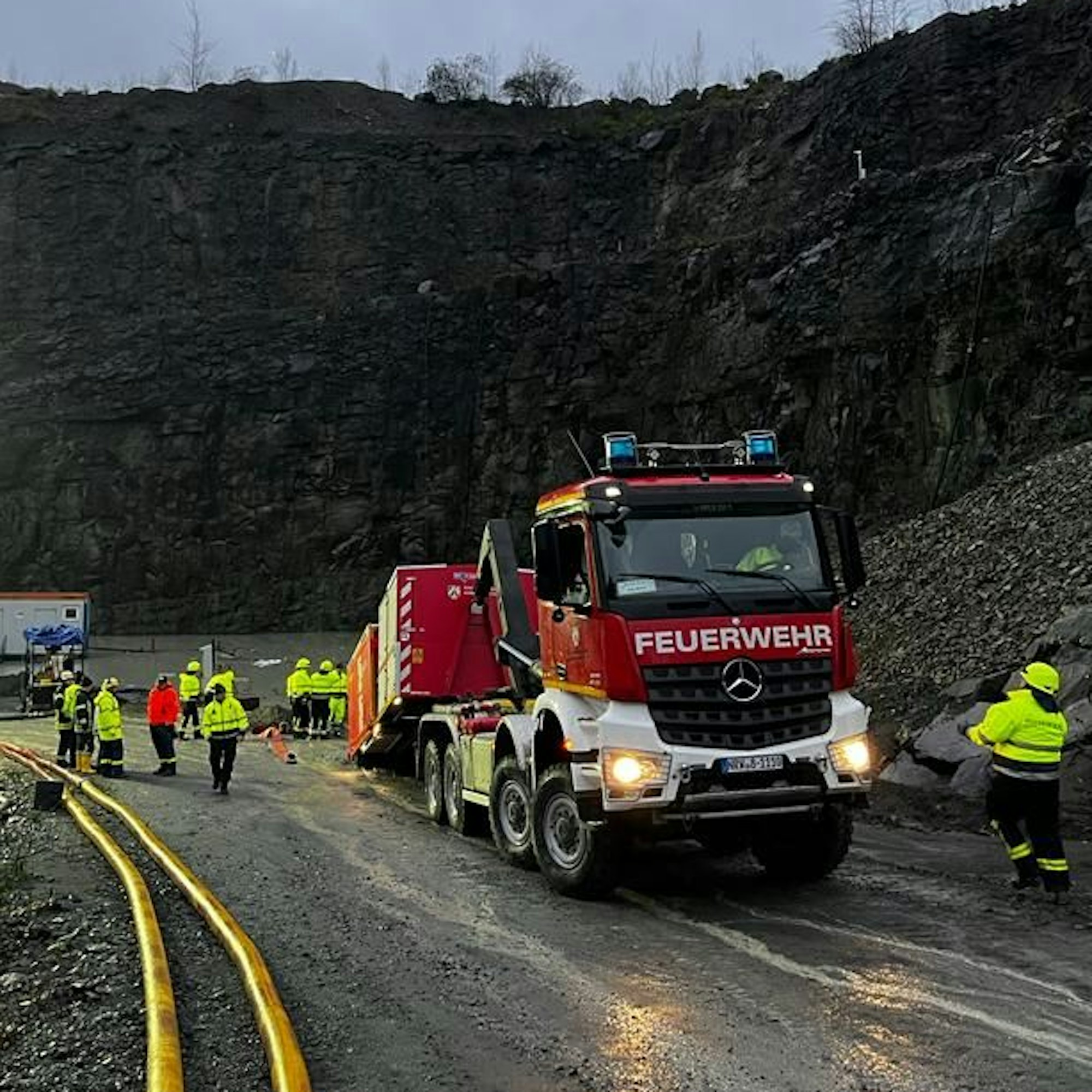 Ein Feuerwehrfahrzeug mit einem Abrollcontainer und Schläuchen steht neben einem gefluteten Steinbruch.
