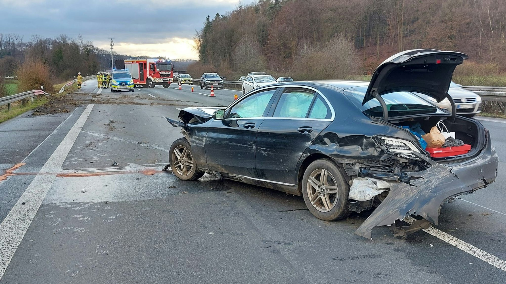 Ein Wagen mit deutlichen Unfallspuren steht auf der Autobahn, die zerdrückte Leitplanke, aufgewühlte Erde und Einsatzfahrzeuge von Feuerwehr und Polizei sind im Hintergrund zu sehen.