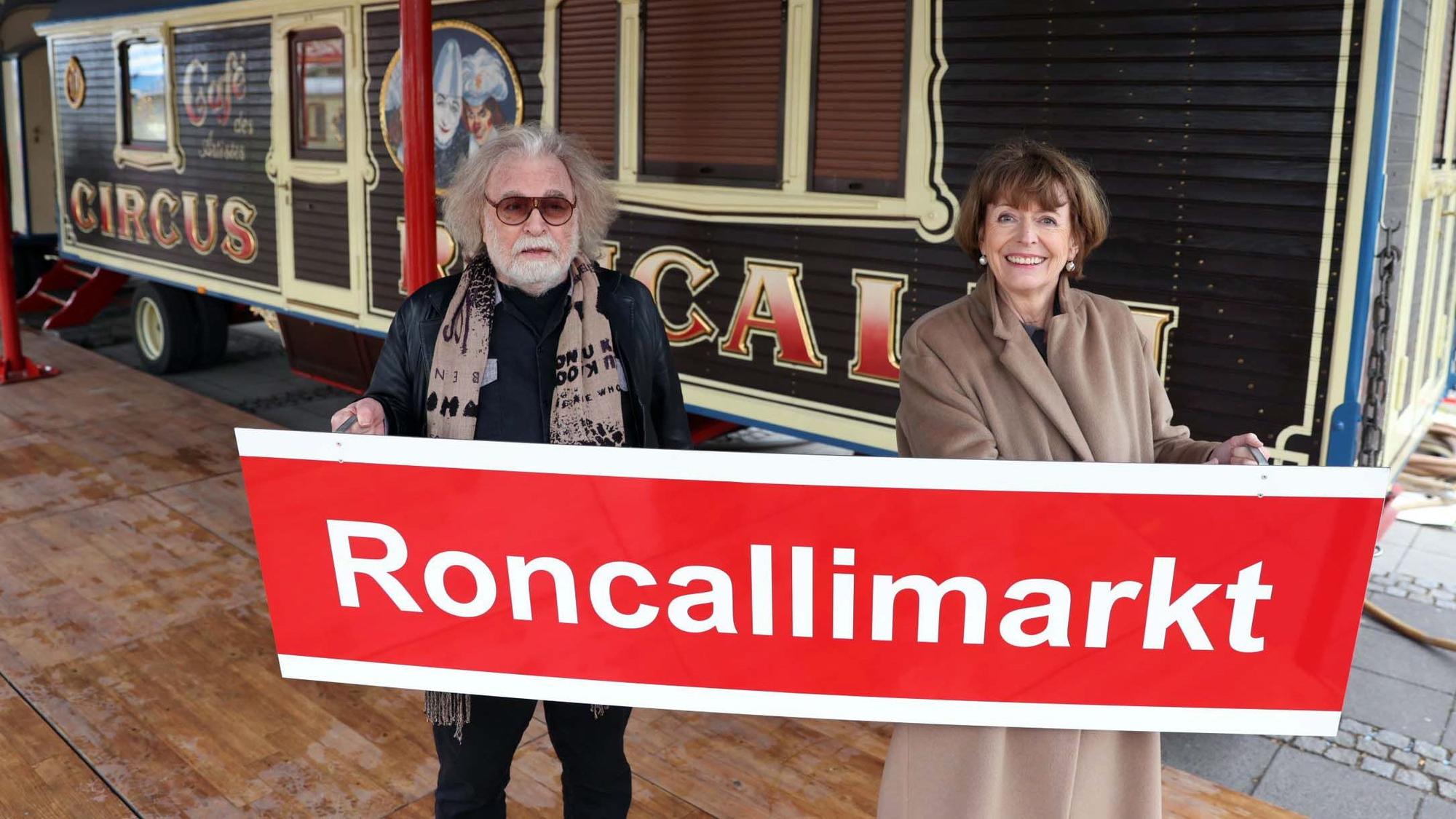 Bernhard Paul und Henriette Reker stehen vor einem Zirkuswagen und halten ein Schild mit der Aufschrift „Roncallimarkt“ hoch.