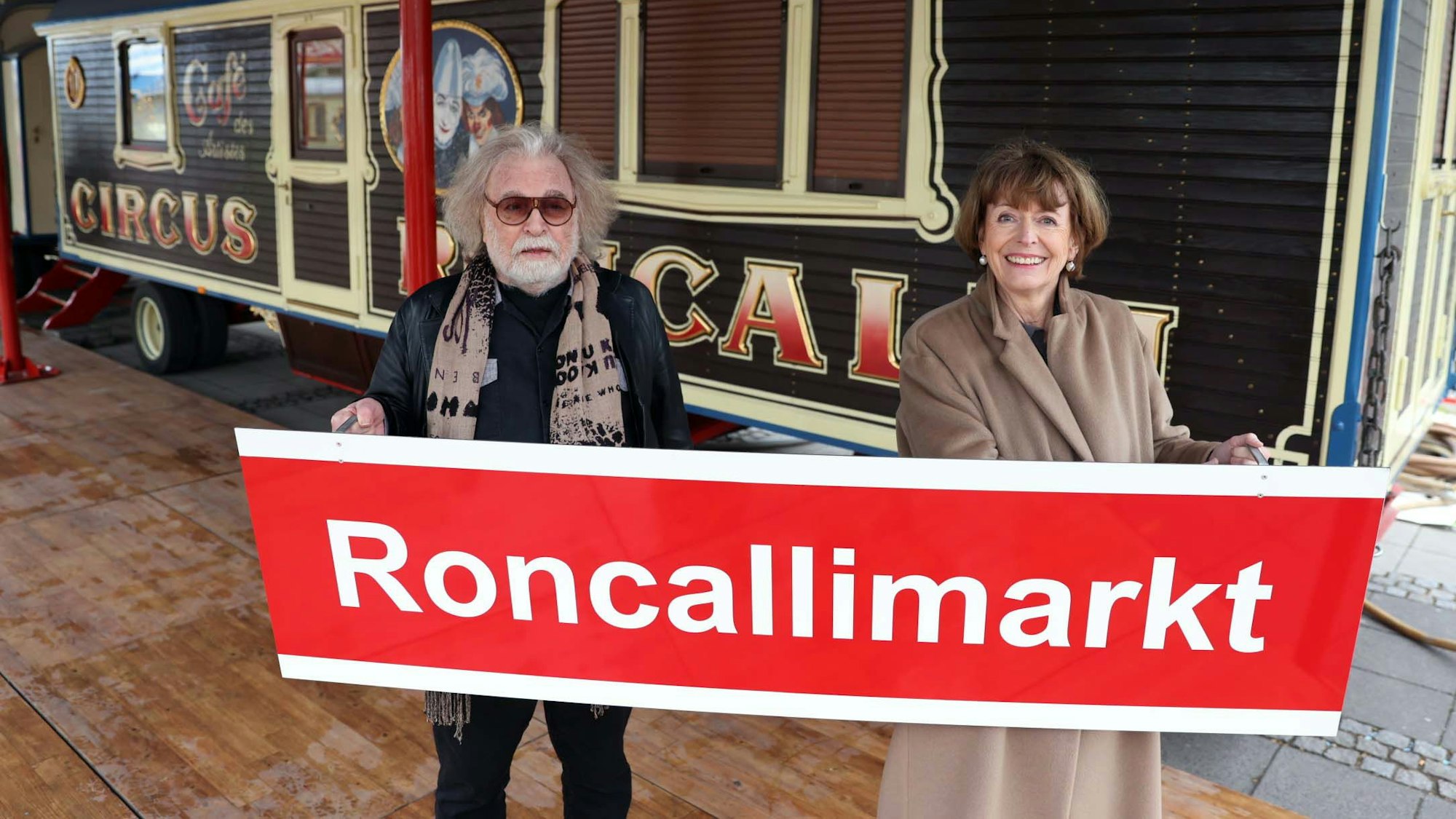 Bernhard Paul und Henriette Reker stehen vor einem Zirkuswagen und halten ein Schild mit der Aufschrift „Roncallimarkt“ hoch.