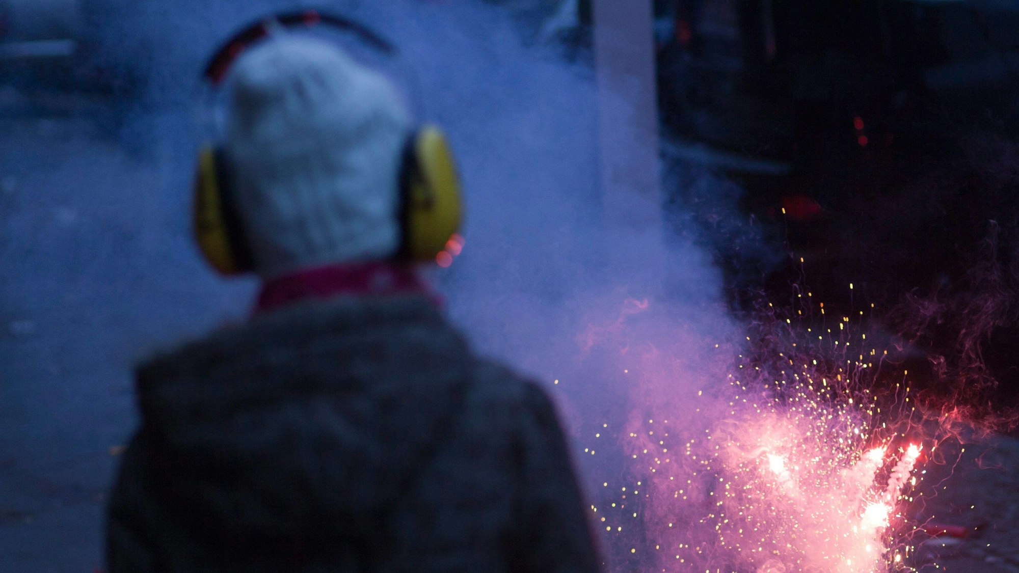 Ein Kind trägt Ohrschützer, es steht in der Nähe eines zündenden Feuerwerkskörpers.
