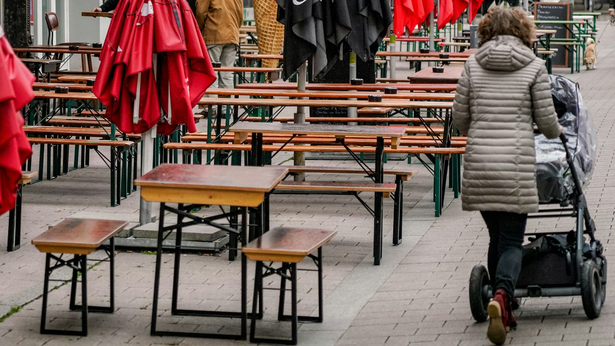 Eine Frau mit einem Kinderwagen ist von hinten zu sehen. Sie geht an unbesetzten Bierzeltgarnituren und zusammengeklappten Sonnenschirmen vorbei.