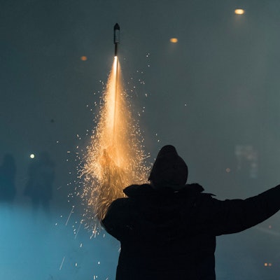Eine Person hält auf einer Straße in der einen Hand eine zündende Rakete, in der anderen einen Böller, im Hintergrund Rauchschwaden von Feuerwerk.