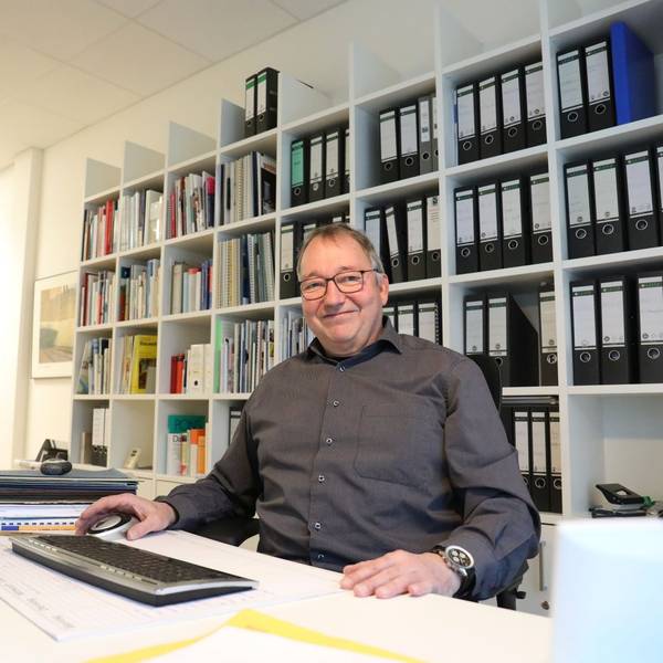 Ein Mann sitzt am Schreibtisch eines Büros, vor sich eine Tastatur. Im Hintergrund steht ein Regal voller Aktenordner.