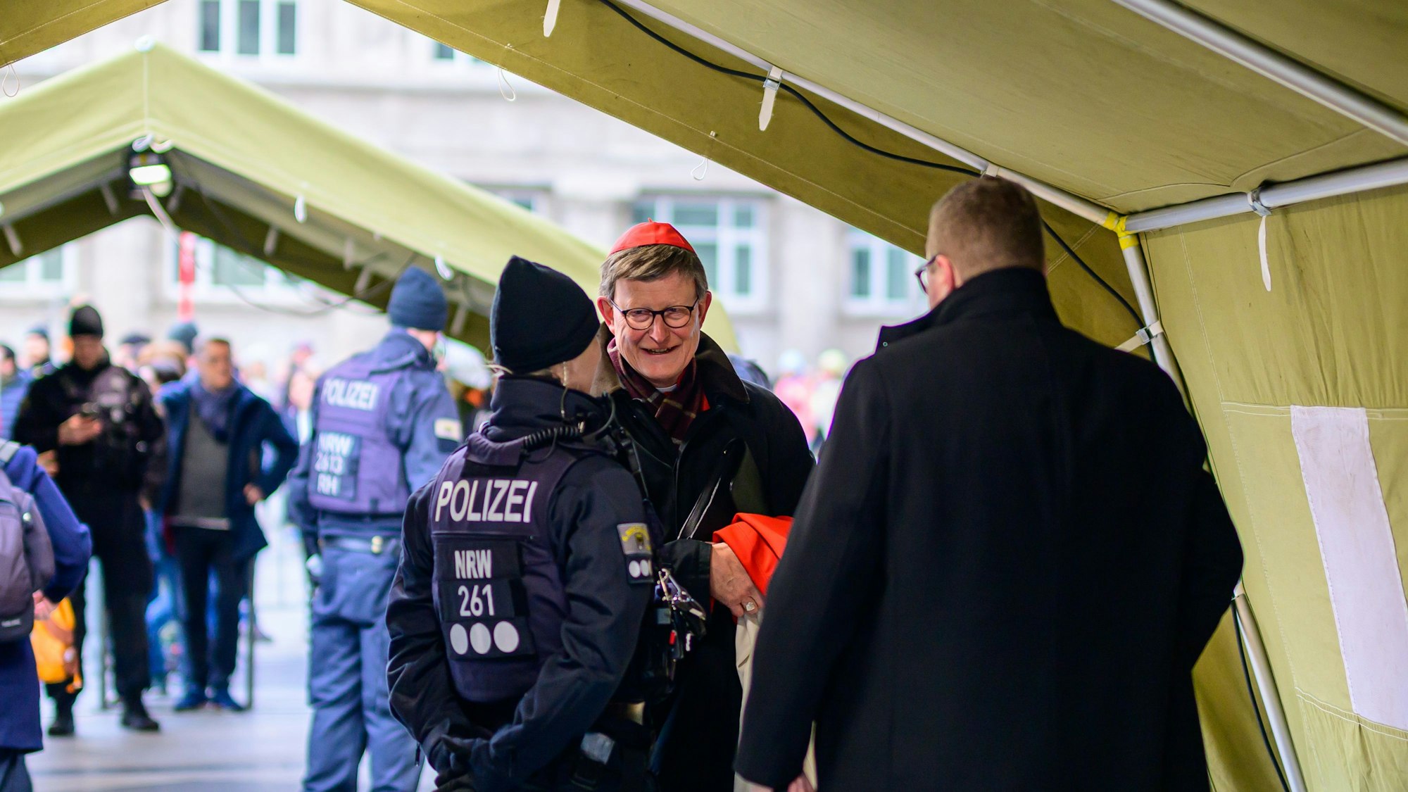Kardinal Woelki geht durch ein Zelt. Eine Beamtin steht vor ihm.