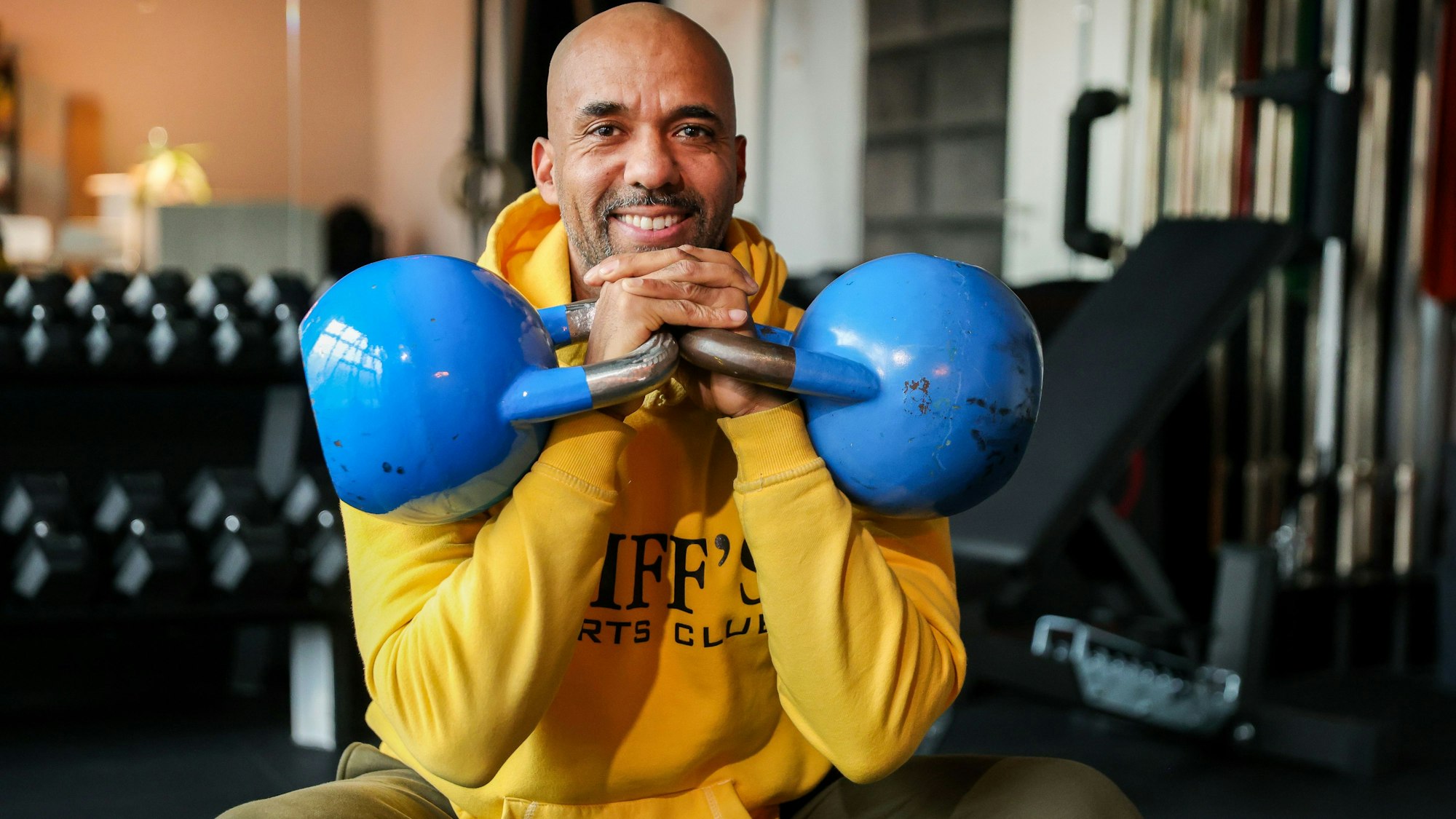 Trainer und Coach Clifford Opoku-Afri sitzt mit zwei blauen Kettlebells auf einer Bank in seinem Fitness-Studio in Ehrenfeld.