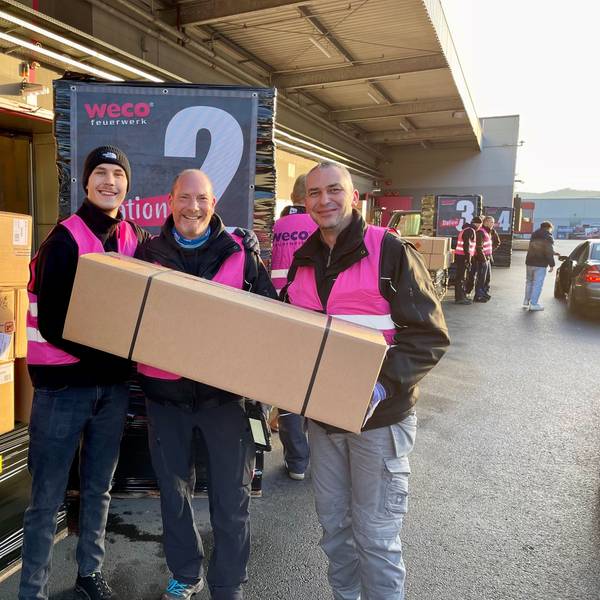 Drei Männer in pinkfarbenen Weco-Westen halten ein langes Überraschungspaket.