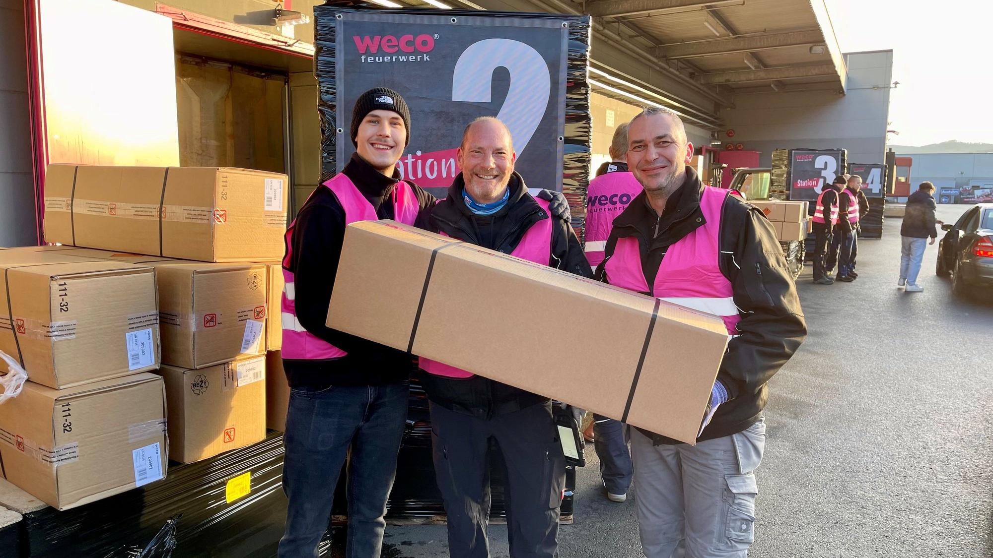 Drei Männer in pinkfarbenen Weco-Westen halten ein langes Überraschungspaket.