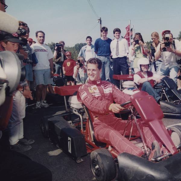 Michael Schumacher sitzt in einem roten Ferrari-Overall auf einem Kart umgeben von Fotografen.