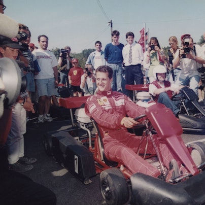 Michael Schumacher sitzt in einem roten Ferrari-Overall auf einem Kart umgeben von Fotografen.