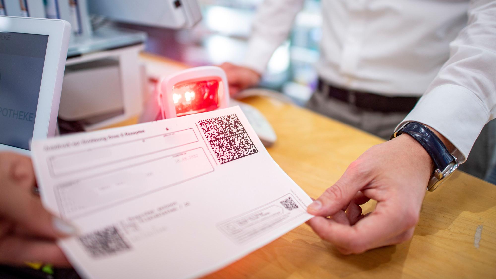 Ein Rezept mit einem QR-Code wird einem Mitarbeiter in einer Apotheke überreicht.