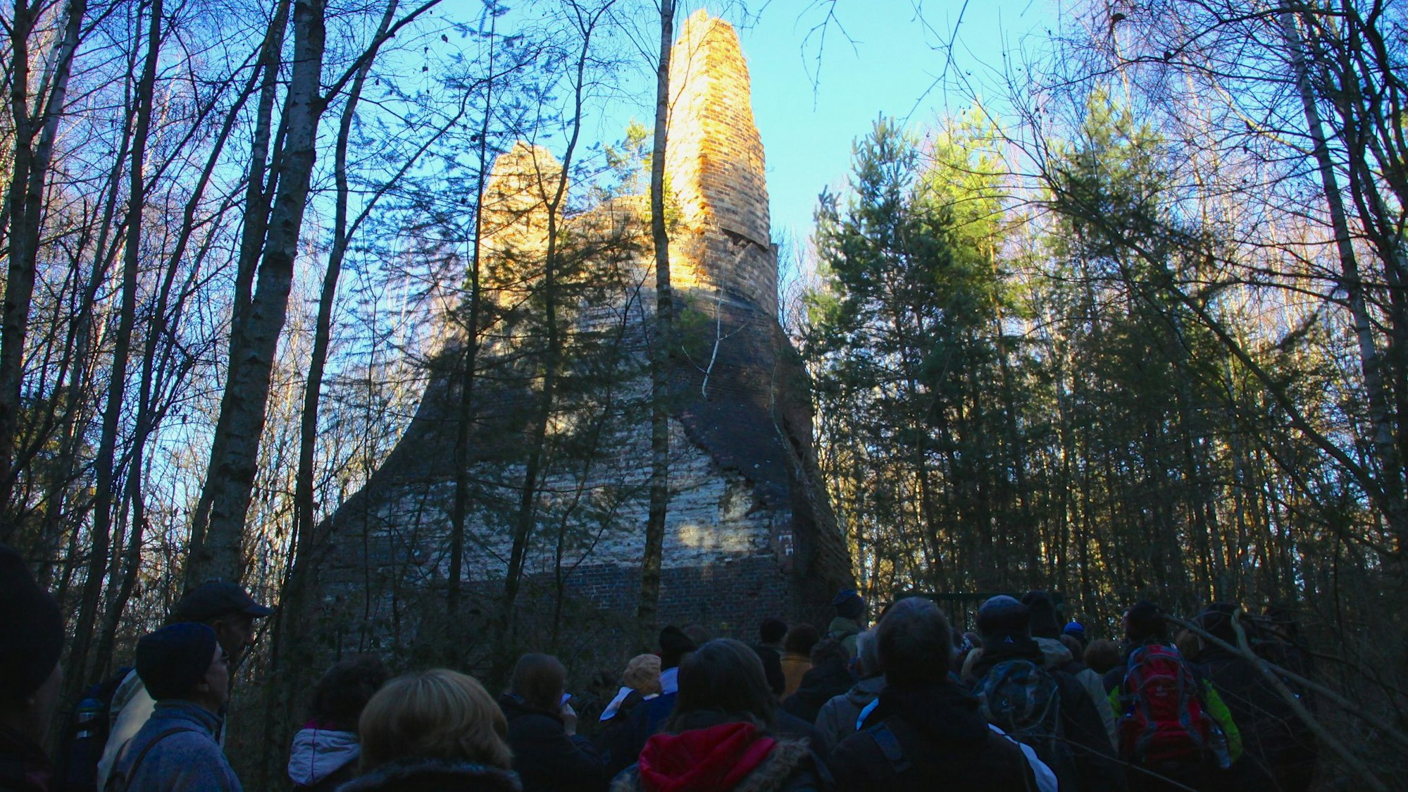 Eine Gruppe Menschen steht im Schatten, umgeben von Bäumen und von der Sonne beschienen ist der Lange Emil, ein Relikt aus der Bergbauzeit in Mechernich.