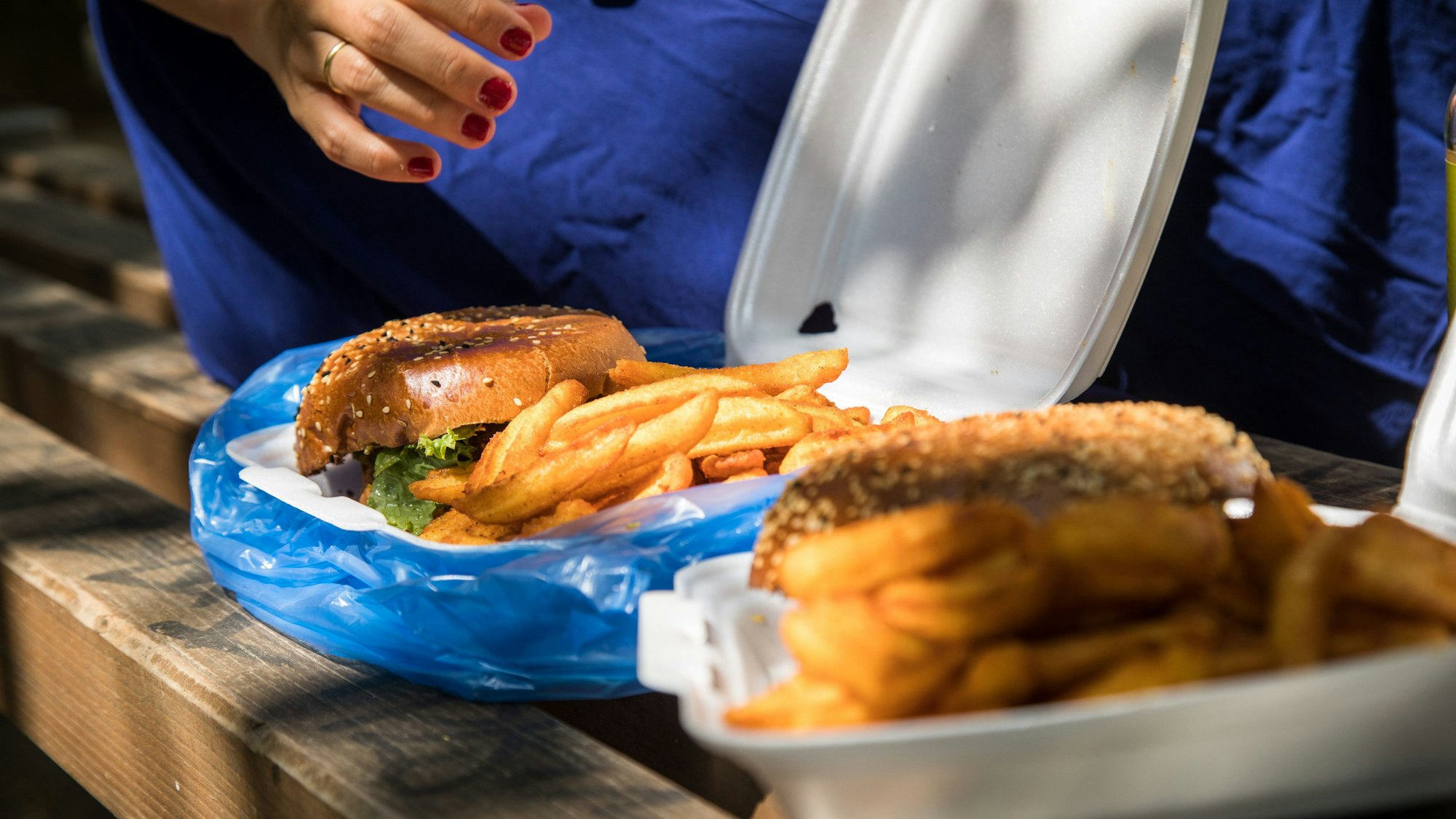 Eine Hand greift auf einer Parkbank in eine Kunststoffverpackung, ansprechend mit Pommes und Burger gefüllt.