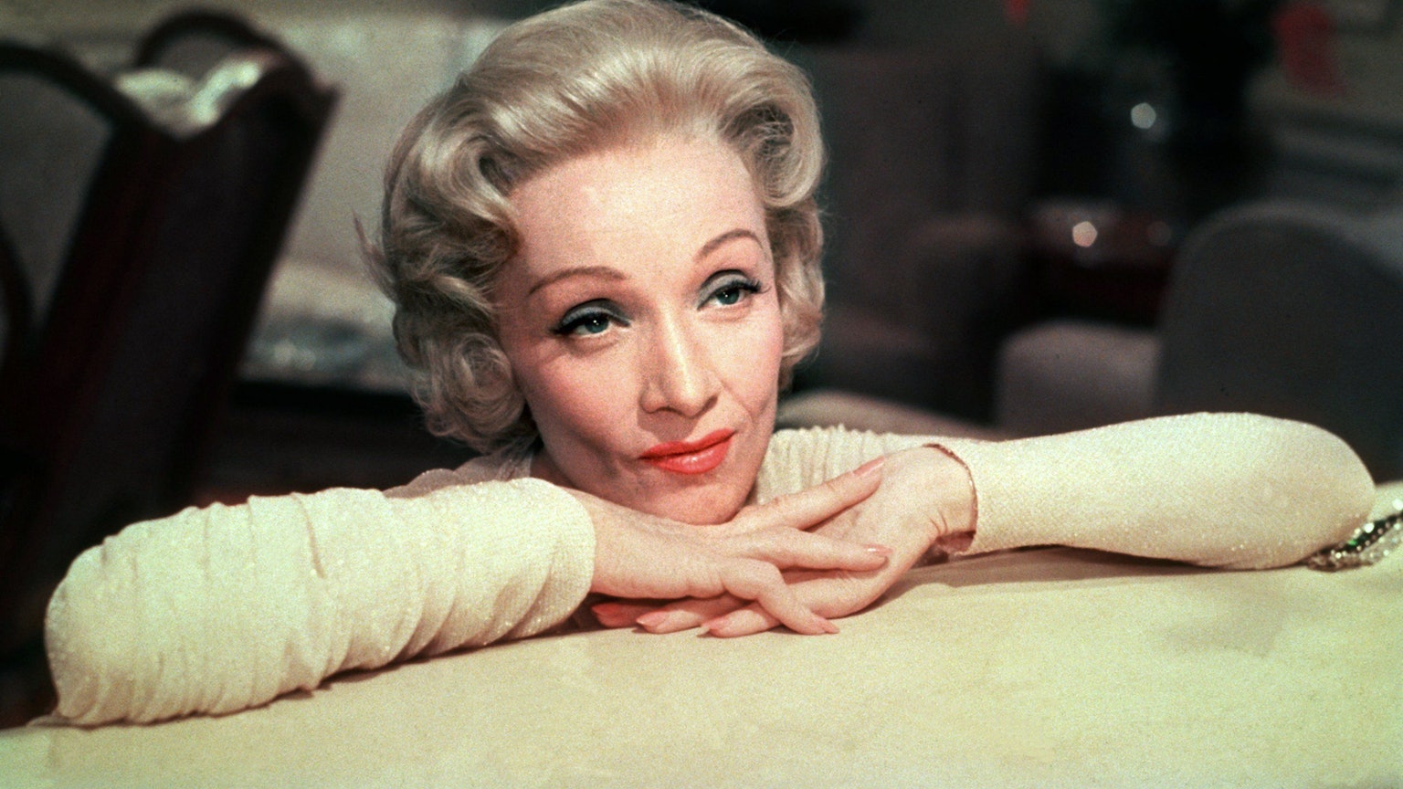 Marlene Dietrich hat ihren Kopf auf ihren verschränkten Händen, die auf einem Tisch liegen, abgelegt.