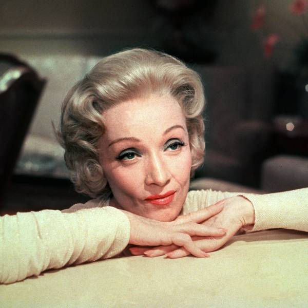 Marlene Dietrich hat ihren Kopf auf ihren verschränkten Händen, die auf einem Tisch liegen, abgelegt.
