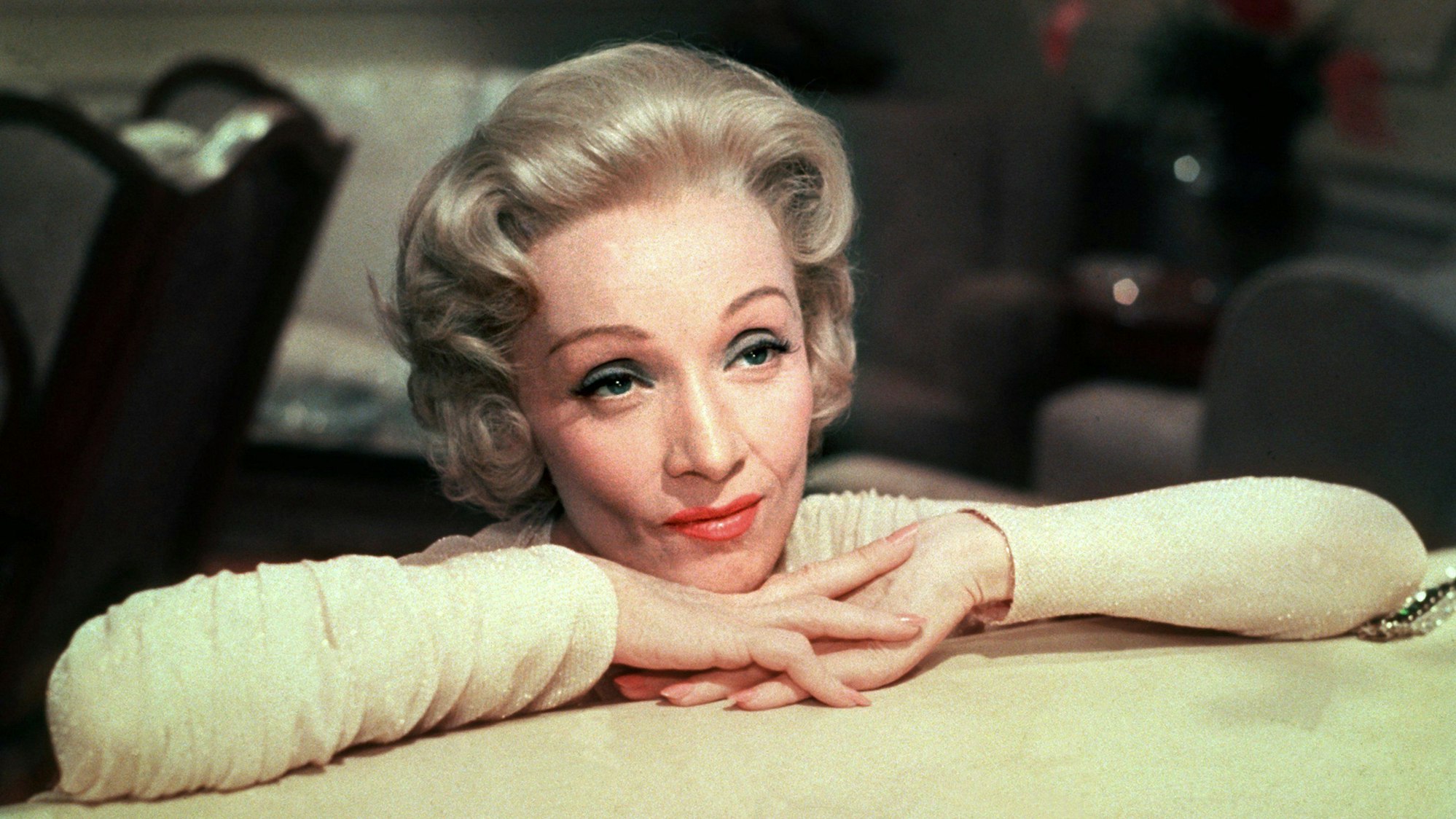 Marlene Dietrich hat ihren Kopf auf ihren verschränkten Händen, die auf einem Tisch liegen, abgelegt.