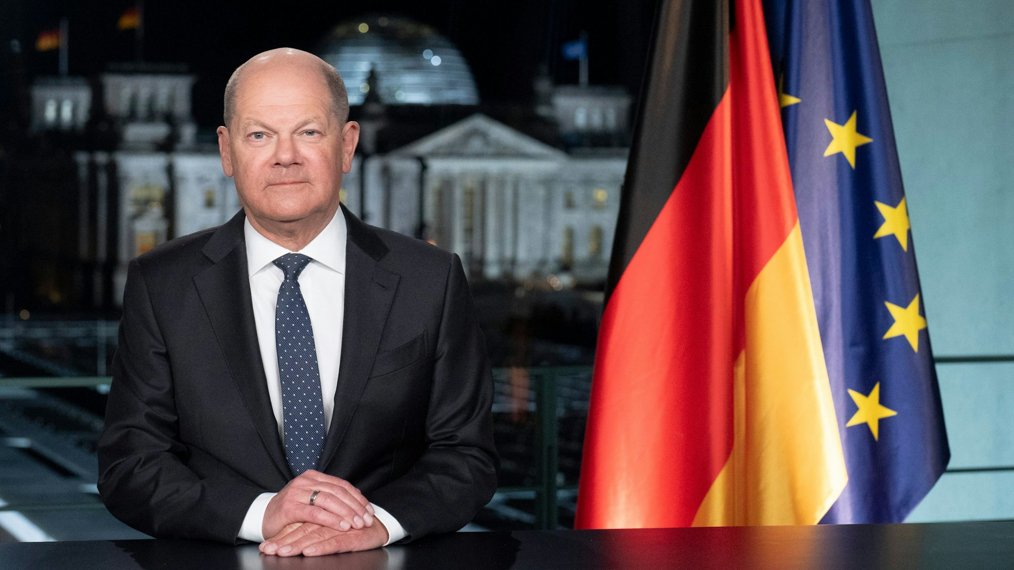 Bundeskanzler Olaf Scholz bei seiner Neujahrsansprache für 2024. Er sitzt an einem dunklen Tisch, neben ihm eine Deutschlandflagge und die Flagge der Europäischen Union.
