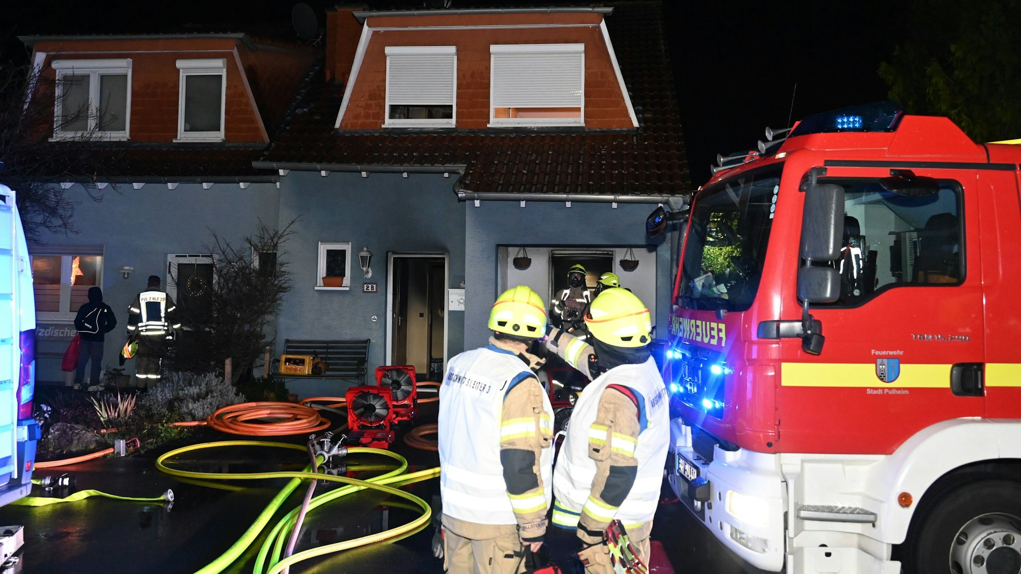 Feuerwehrleute und ein Einsatzwagen der Feuerwehr stehen vor einem Haus, das gebrannt hat. Im Vorgarten liegen Schläuche.