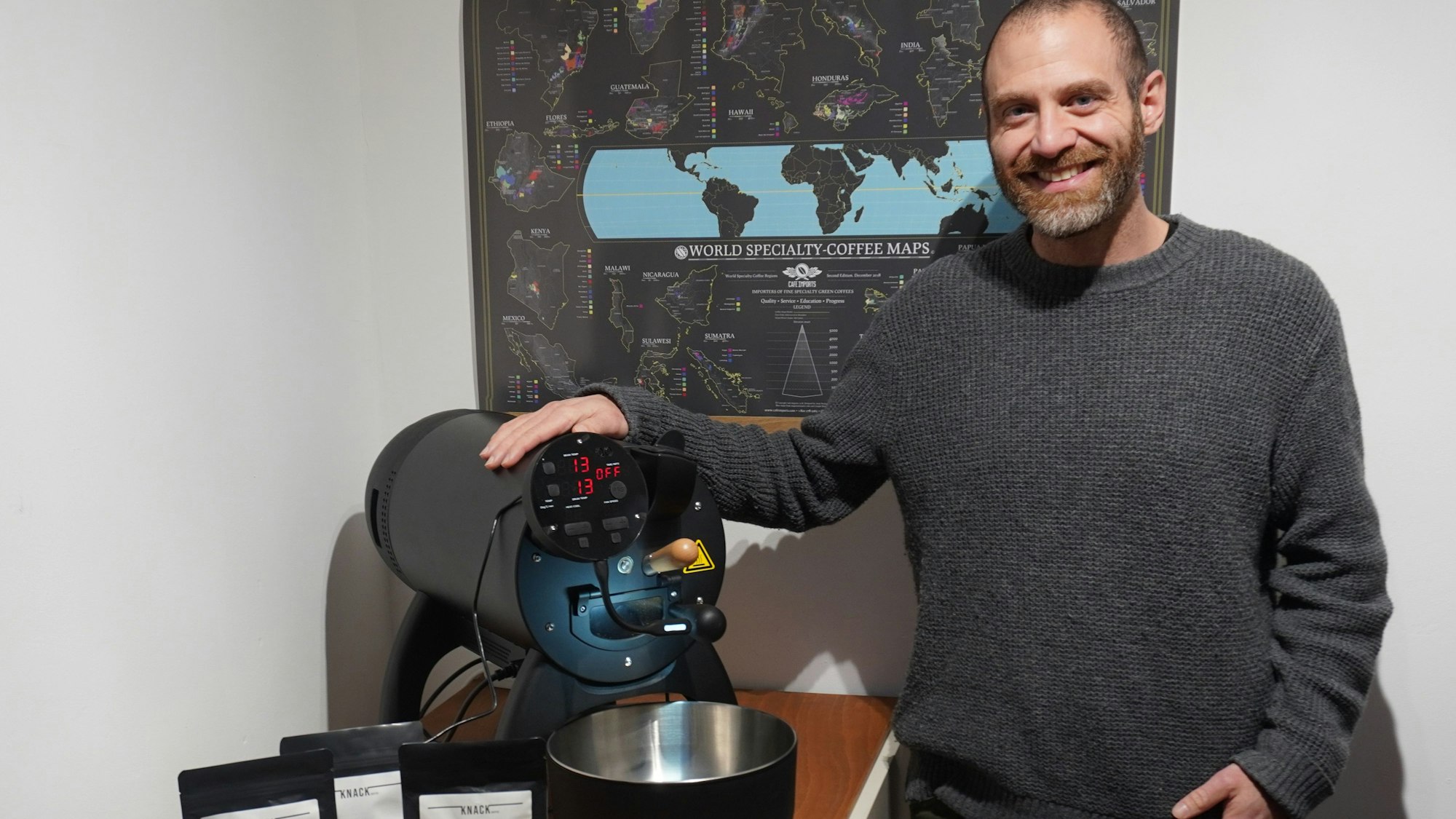 Ein Mann lächelt in die Kamera. Neben ihm auf einem Tisch steht eine Kaffeeröstmaschine.