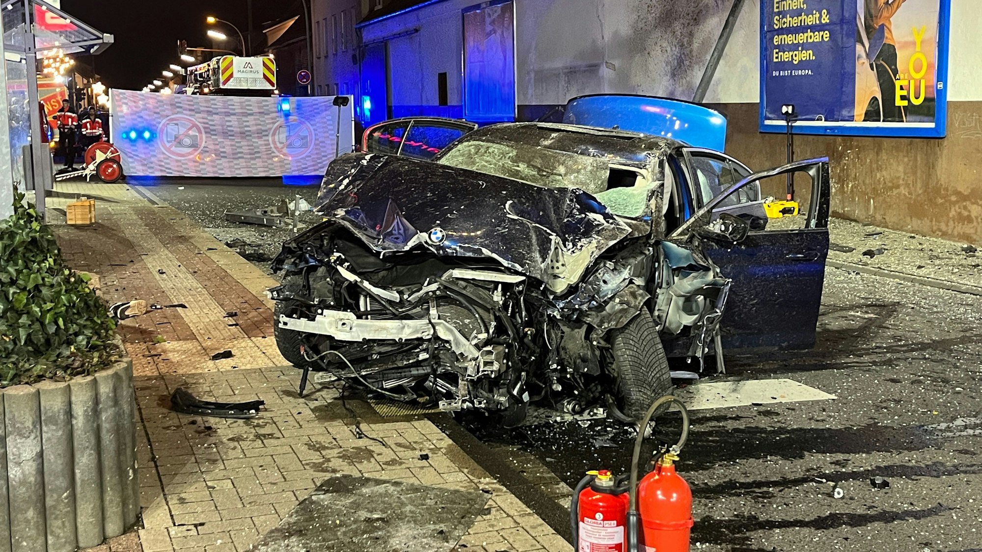Auf der nächtlichen Provinzialstraße in Niederkassel-Mondorf steht das völlig zerstörte Wrack eine BMW, der in eine Bushaltestelle gerast war.