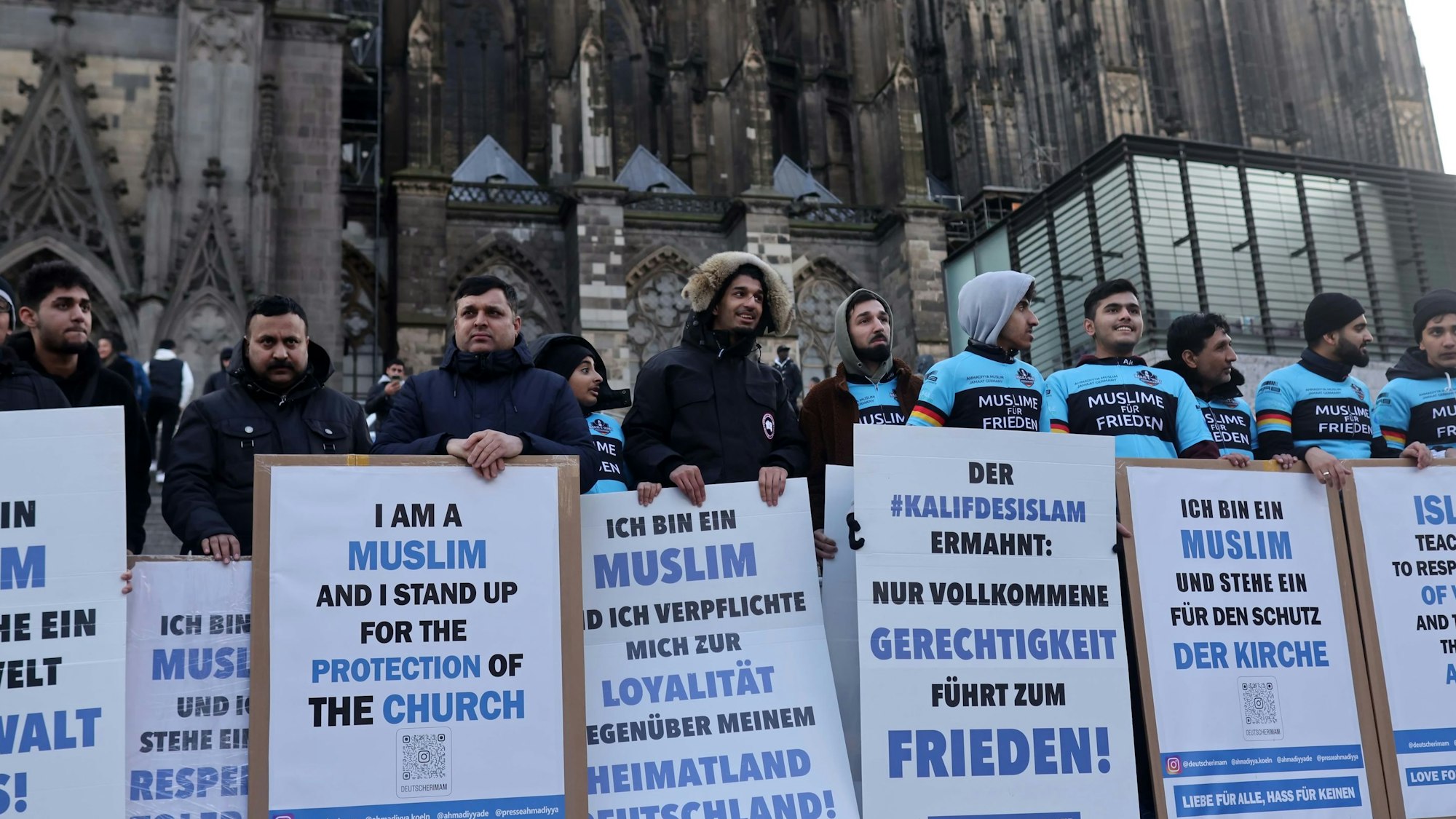 Mitgliederder Ahmadiyya Muslim Gemeinde stehen mit Plakaten, die zu Frieden aufrufen, vor dem Kölner Dom.