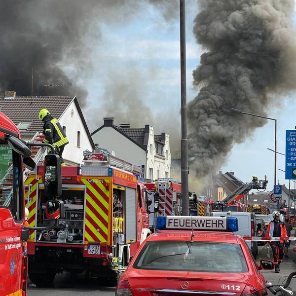 Viele Feuerwehrfahrzeuge stehen ij einer Straße, schwarzer Qualm dringt aus einem Gebäude.
