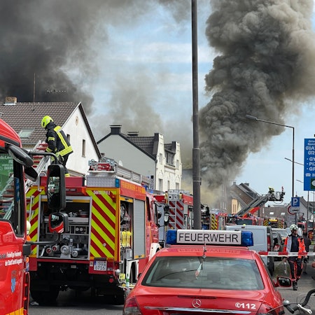 Viele Feuerwehrfahrzeuge stehen ij einer Straße, schwarzer Qualm dringt aus einem Gebäude.