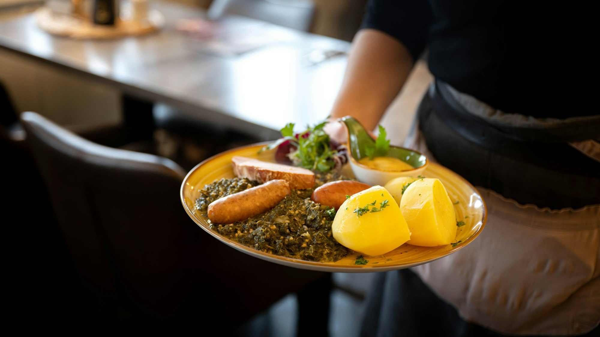 Man sieht den Arm einer Kellnerin, die einen Teller mit Möhren, Kartoffeln, Gehackten und Salat transportiert.