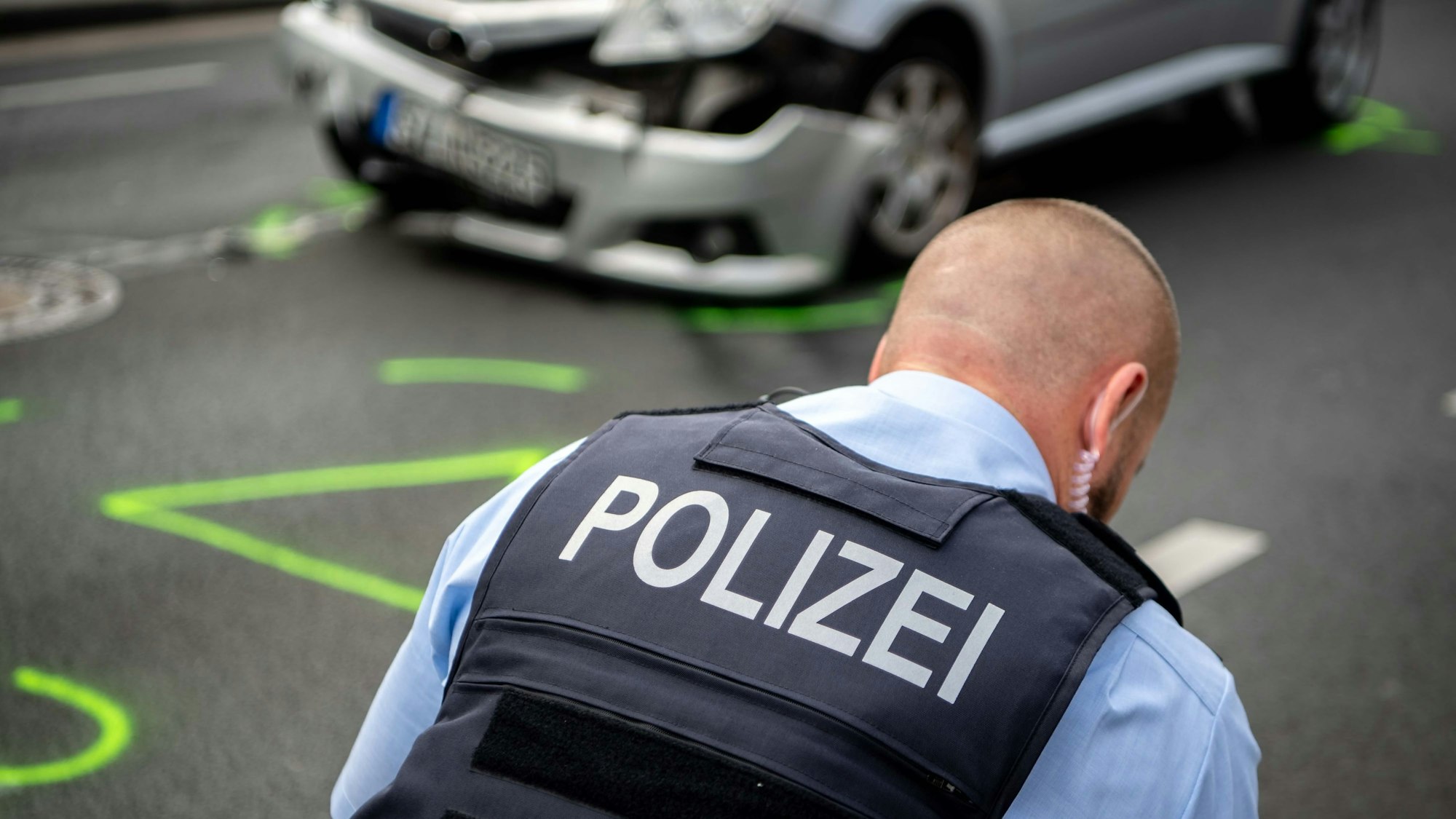 Ein Polizist nimmt einen Verkehrsunfall auf. Im Hintergrund steht ein beschädigtes Auto, auf der Straße wurden Markierungen mit Leuchtfarbe aufgesprüht.