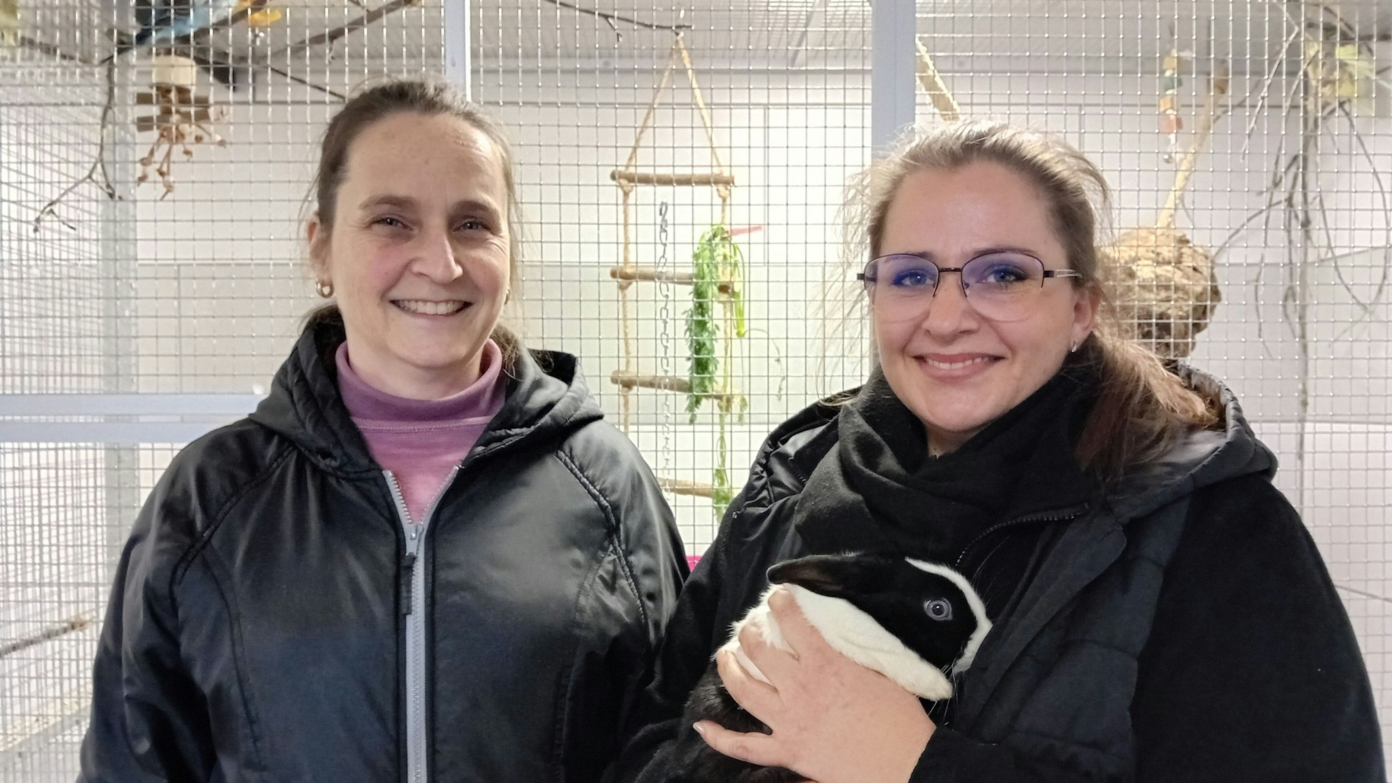 Zwei Frauen vor einer Voliere, die rechte hält ein Kaninchen auf dem Arm.